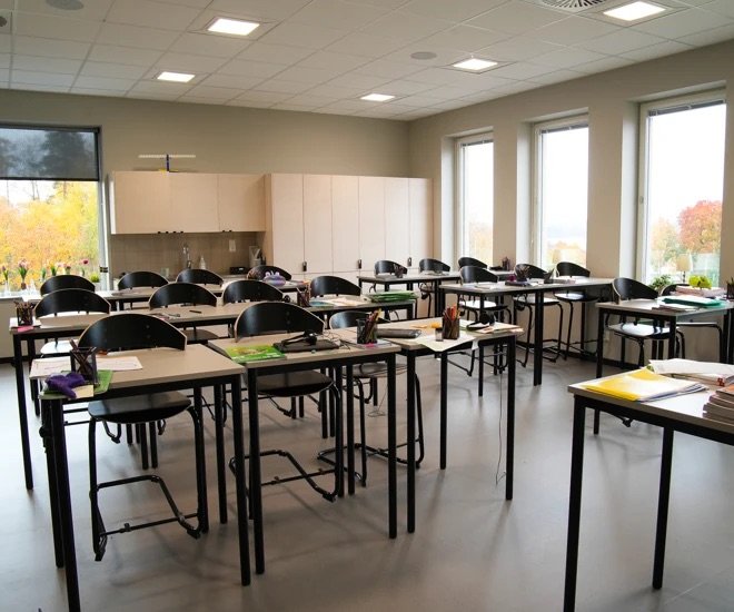 Ett klassrum med tomma skrivbord och stolar, fönster med utsikt över träd, skolmaterial på borden.
