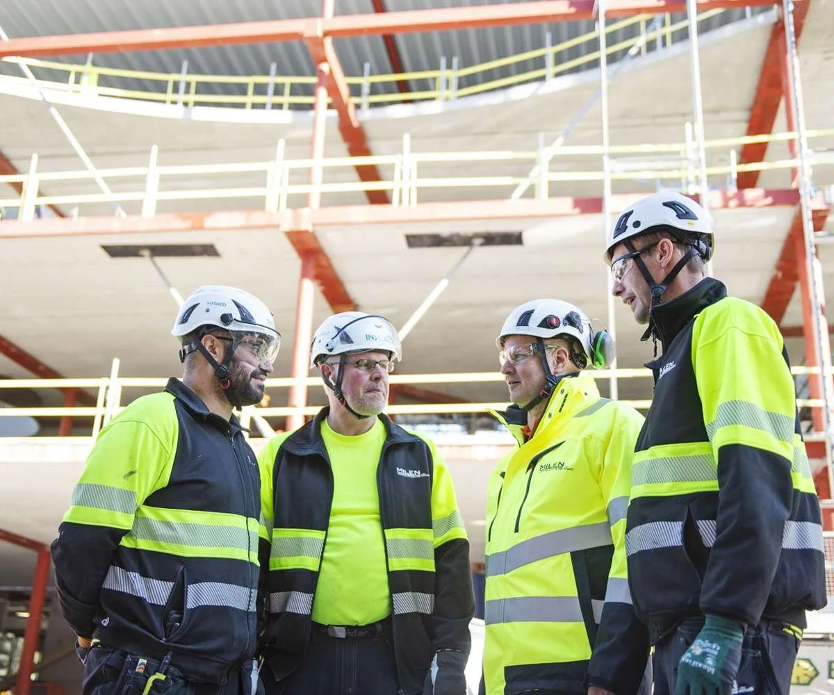 Fyra byggarbetare i skyddsmössor och reflexvästar i samtal på en byggarbetsplats.
