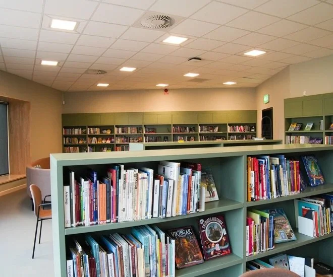 Bokhyllor i ett bibliotek fyllt med böcker, med sittplatser och ett fönster i bakgrunden.