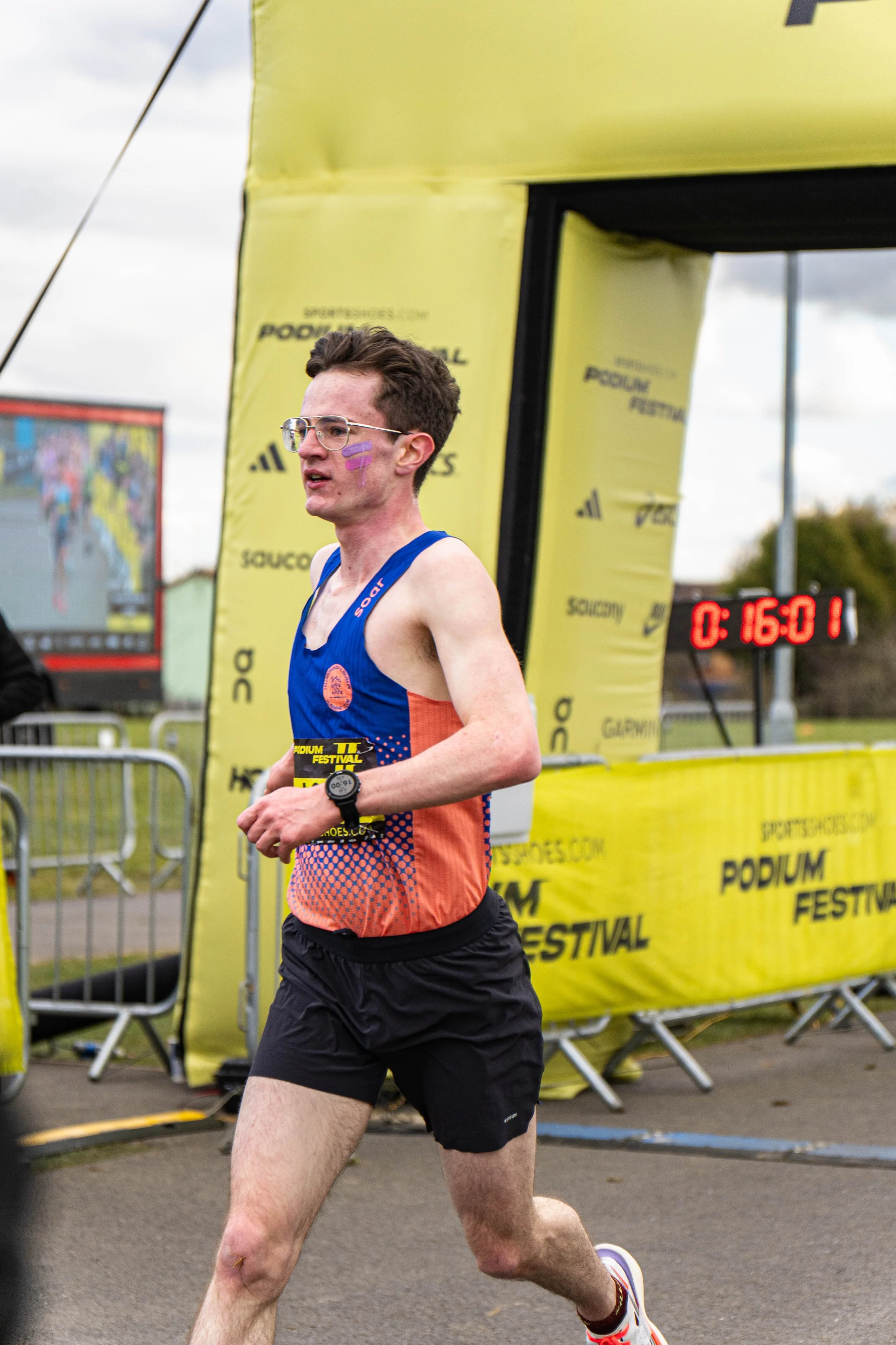 A male runner wearing glasses and a blue and coral race vest, participating in a race, crossing the finish line with a digital timer showing 16 minutes and 1 seconds in the background.