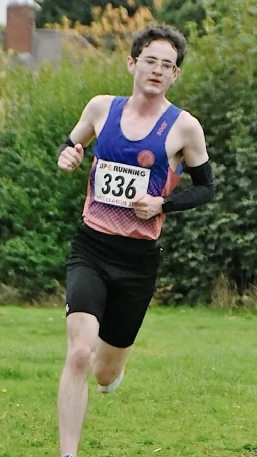 A young man running outdoors on a grassy field during a race, wearing a blue and coral racing vest, black shorts, glasses, and a race bib number 336.