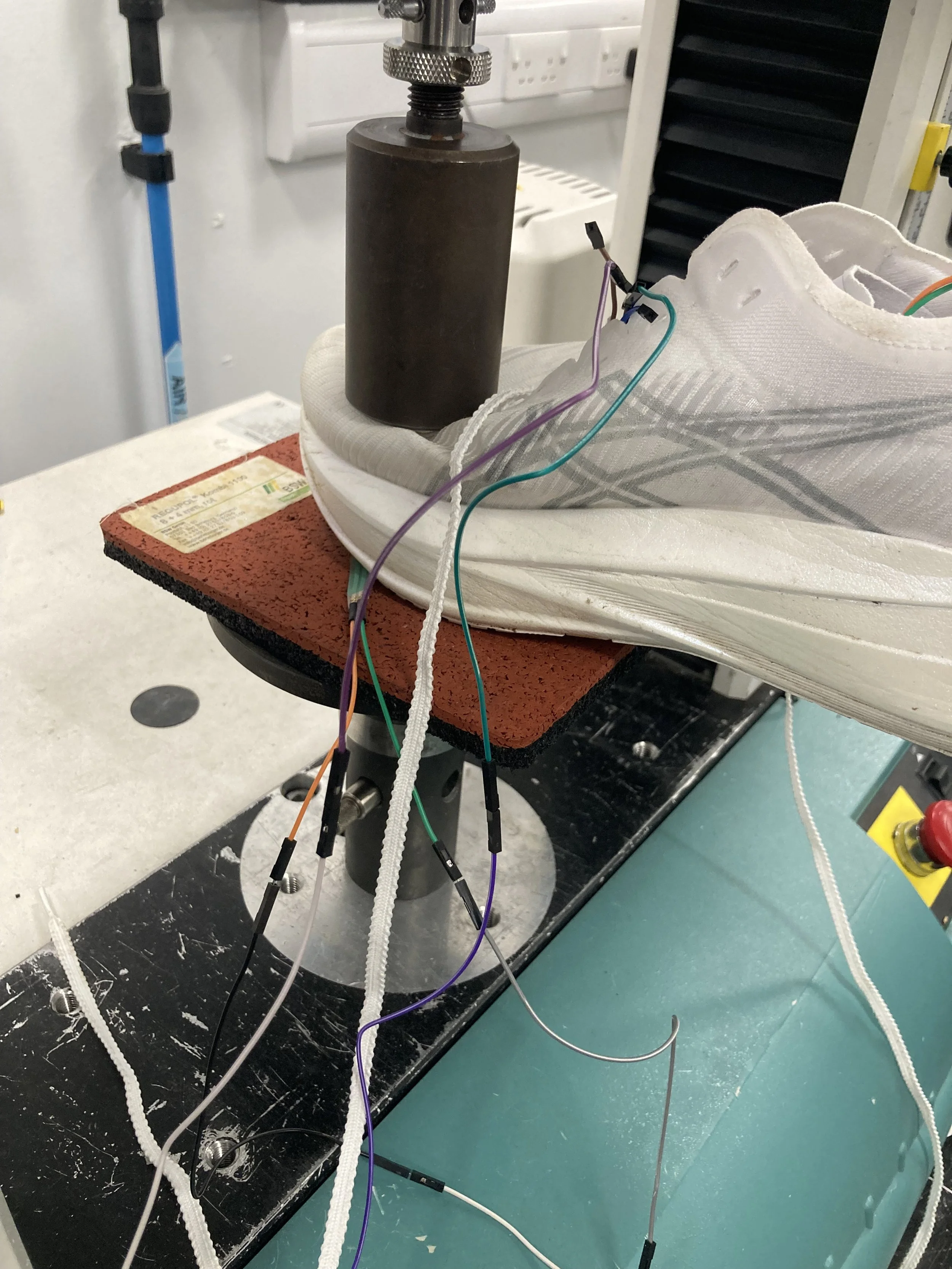 INSTRON compression device consisting of a metal cylinder attachment applying pressure to a white running shoe with wires coming out of the shoe.