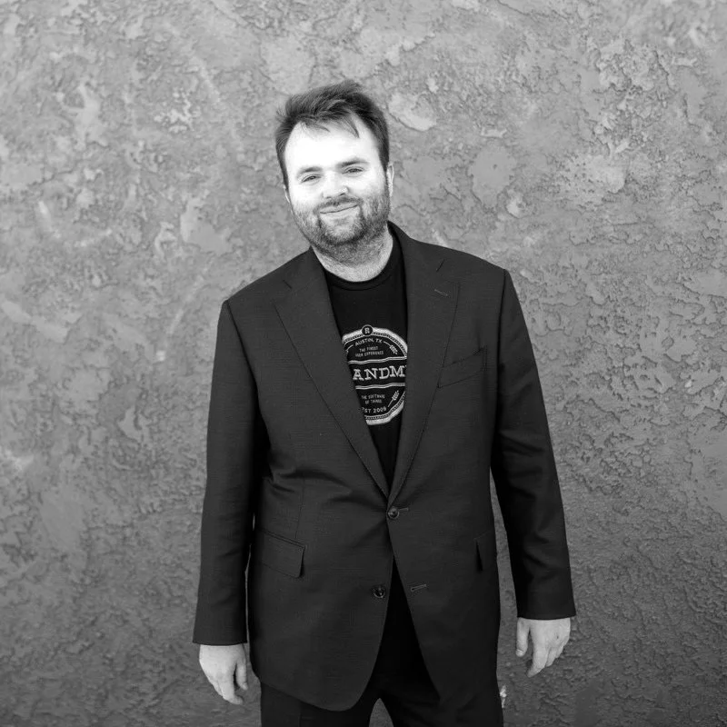 A man with a beard smiling, wearing a black blazer over a black T-shirt, standing against a textured wall.