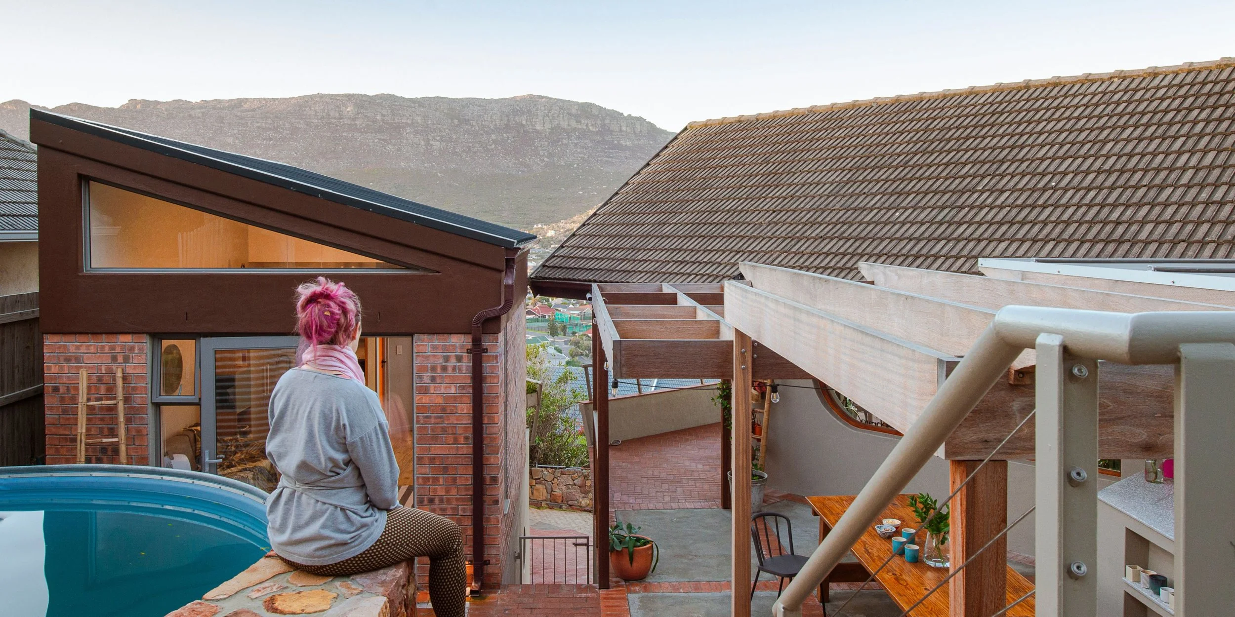 Woman with pink hair sitting on a ledge in an outdoor backyard with a hot tub, wooden pergola, and mountain view in the background.