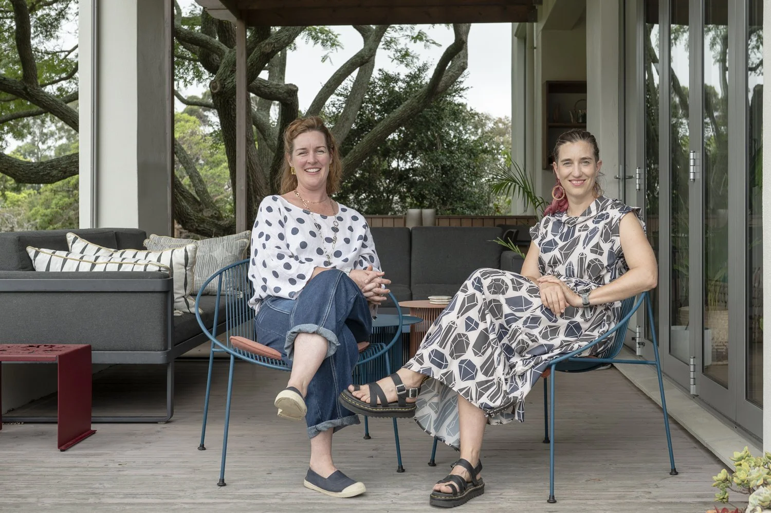 Two women sitting outdoors on a porch, smiling at the camera. One is wearing a polka dot blouse and jeans, the other a patterned dress, with trees visible in the background.