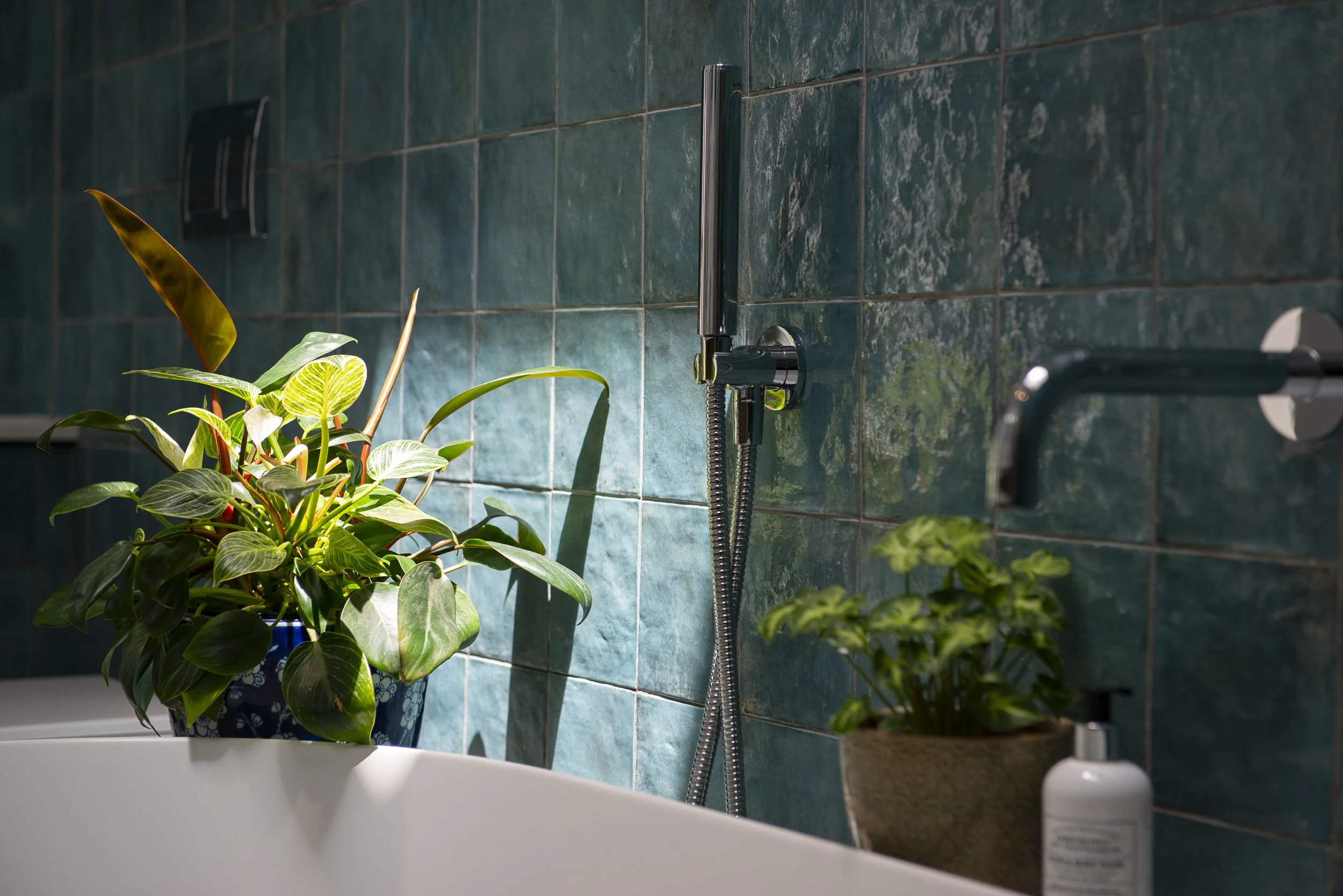 Green tiled bathroom wall with a mounted silver showerhead and a flexible hose. Green plants in ceramic and plastic pots sit on a white sink in the foreground. A white soap dispenser is partially visible on the right.