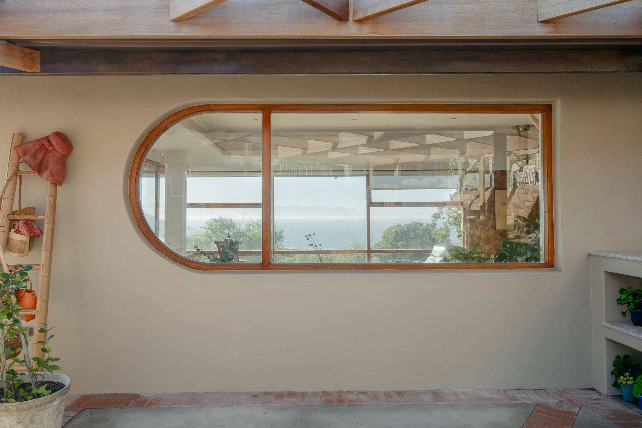 Interior view through a large, oval wooden-framed window looking out onto a balcony with a scenic landscape and water in the distance; part of a house interior with plants and decorative items.