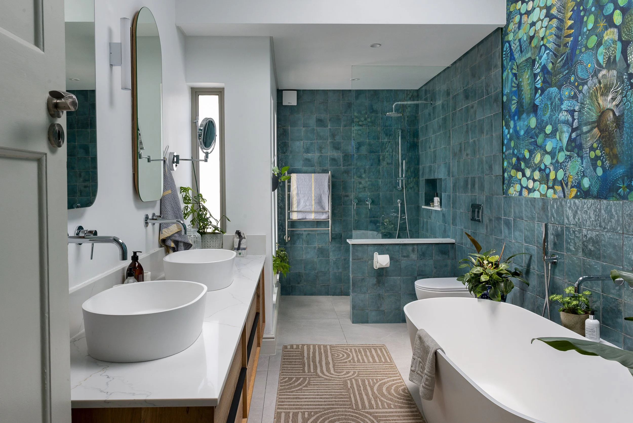 Modern bathroom with white countertops, two vessel sinks, large mirror, plants, teal tile walls, and a bathtub with a shower. There are towels, soap, and a decorative rug on the floor.