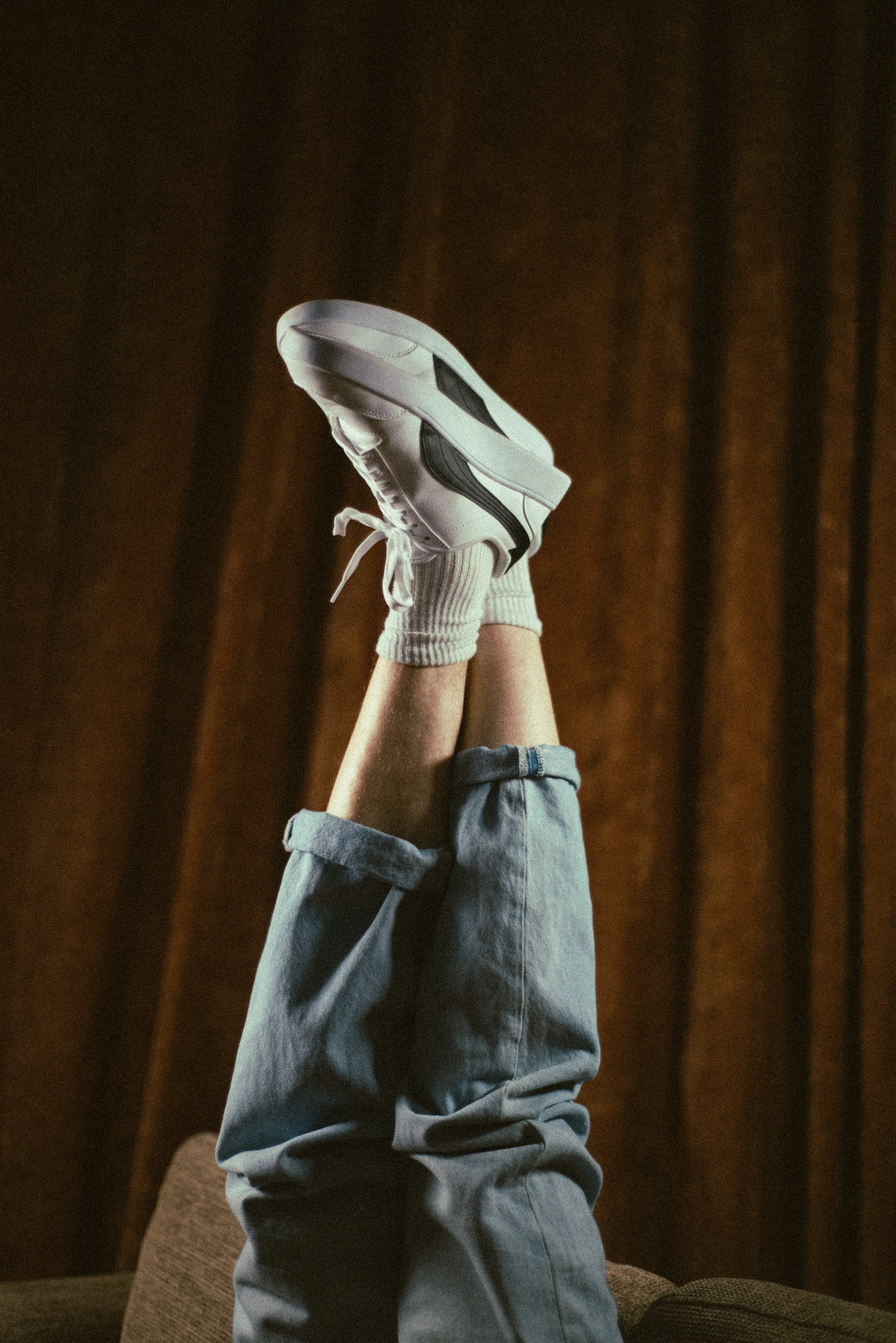 Person wearing white sneakers, gray sock, and loose gray jeans, lifting their legs upward against a dark brown curtain background.