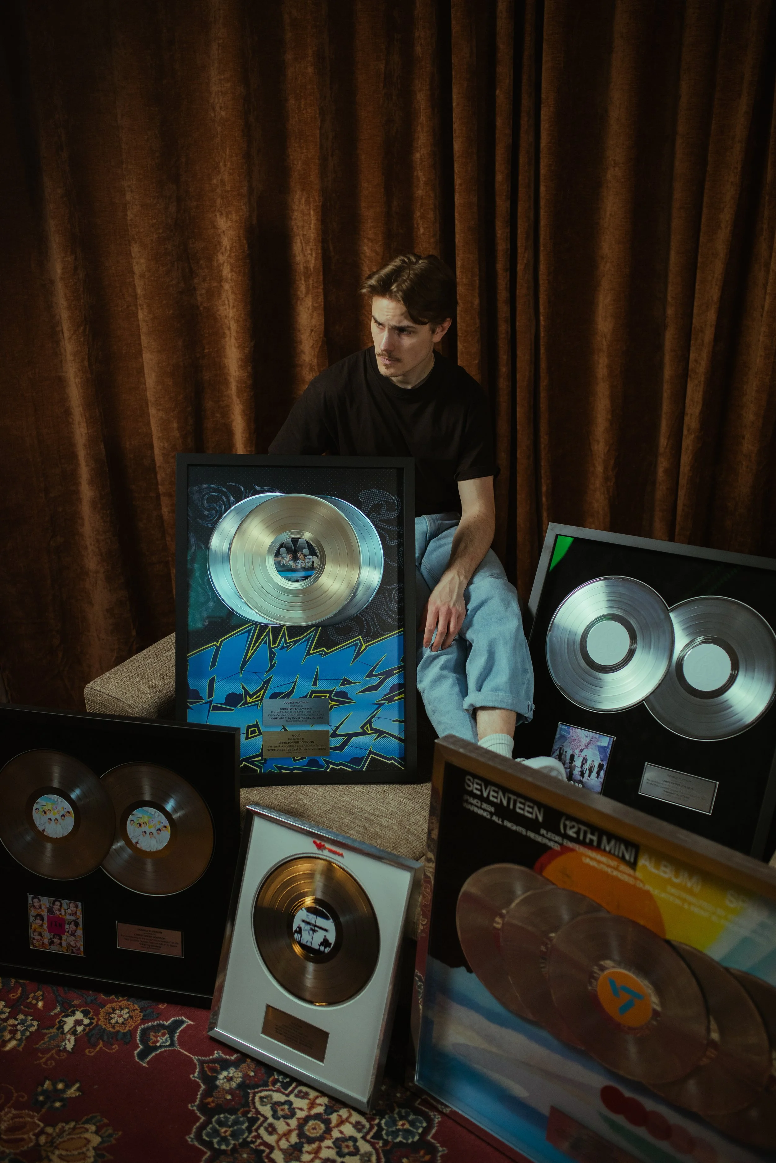 En ung man sitter bland guldskivor och album, med en brun gardin i bakgrunden.