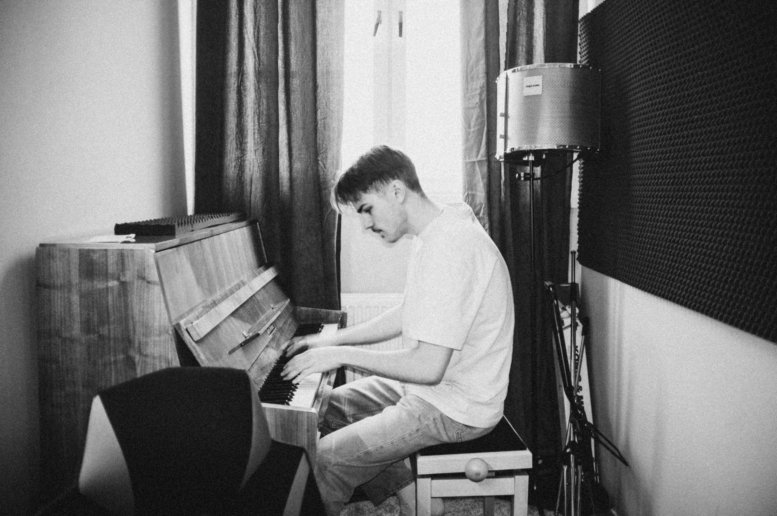 A young man playing a piano in a recording studio with soundproofing panels on the wall and a microphone on a stand nearby.