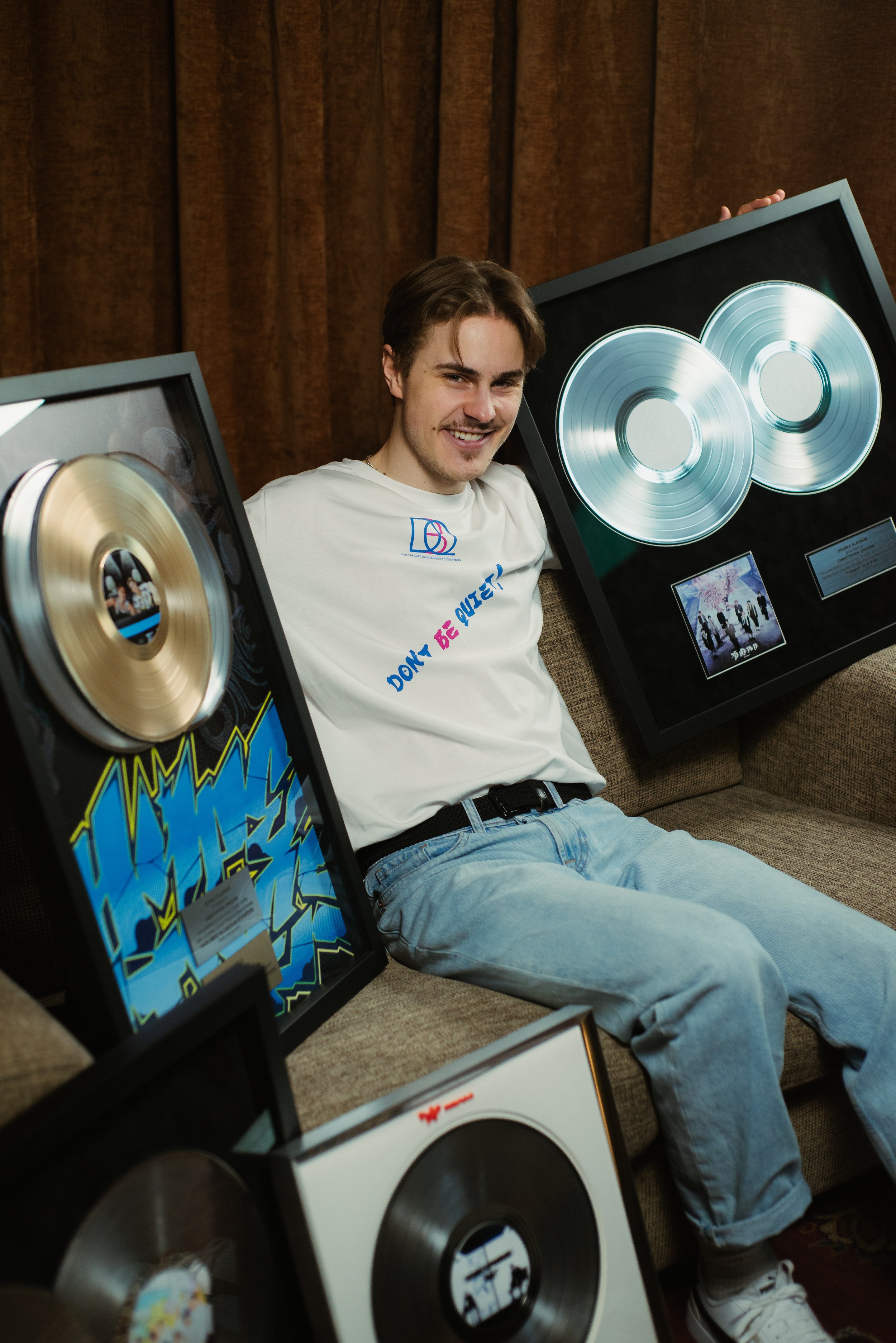 A young man with brown hair and a goatee smiling and sitting on a couch in front of framed album covers, with a brown curtain in the background.
