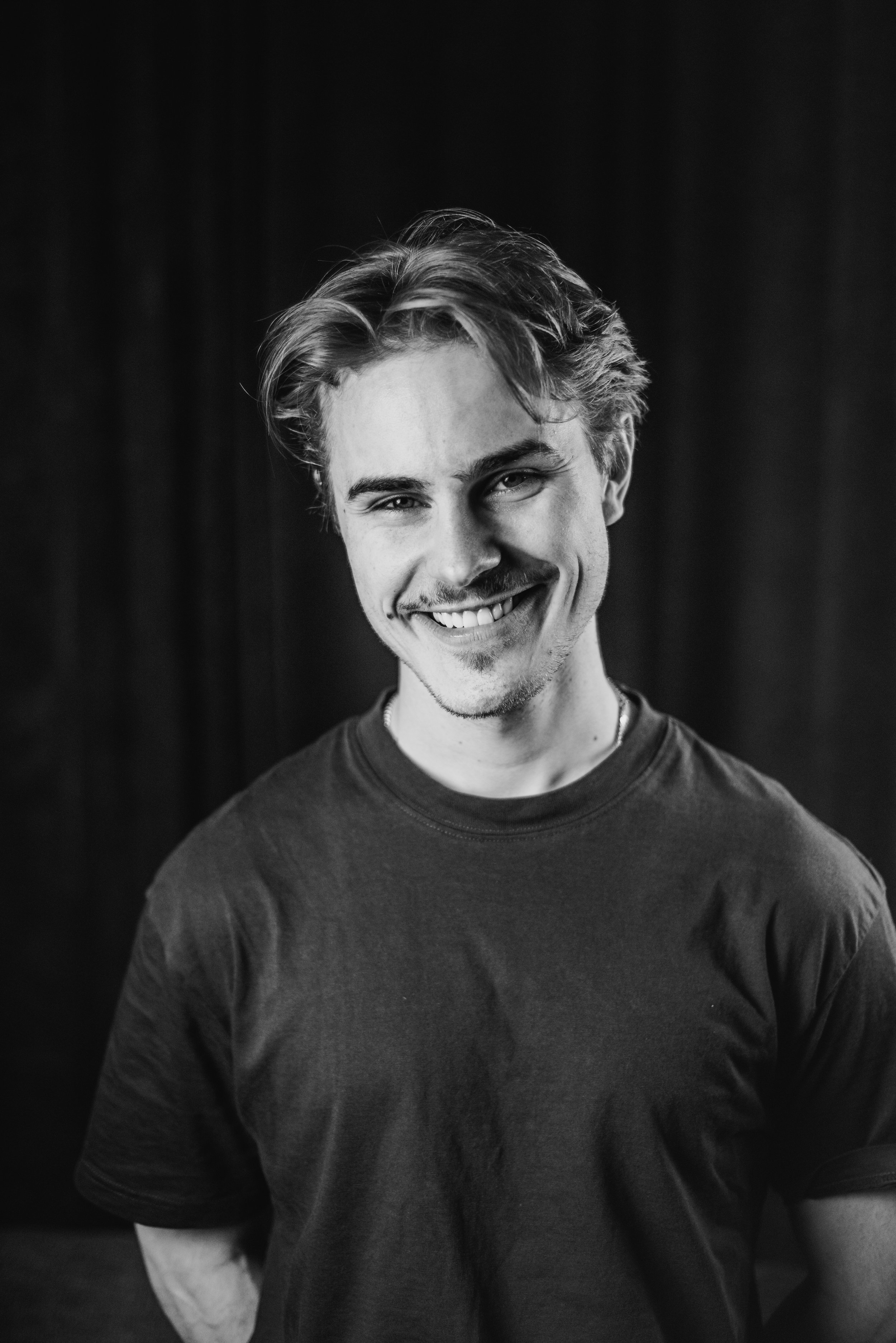 Black and white photo of a smiling young man with wavy hair wearing a t-shirt, standing against a dark background.