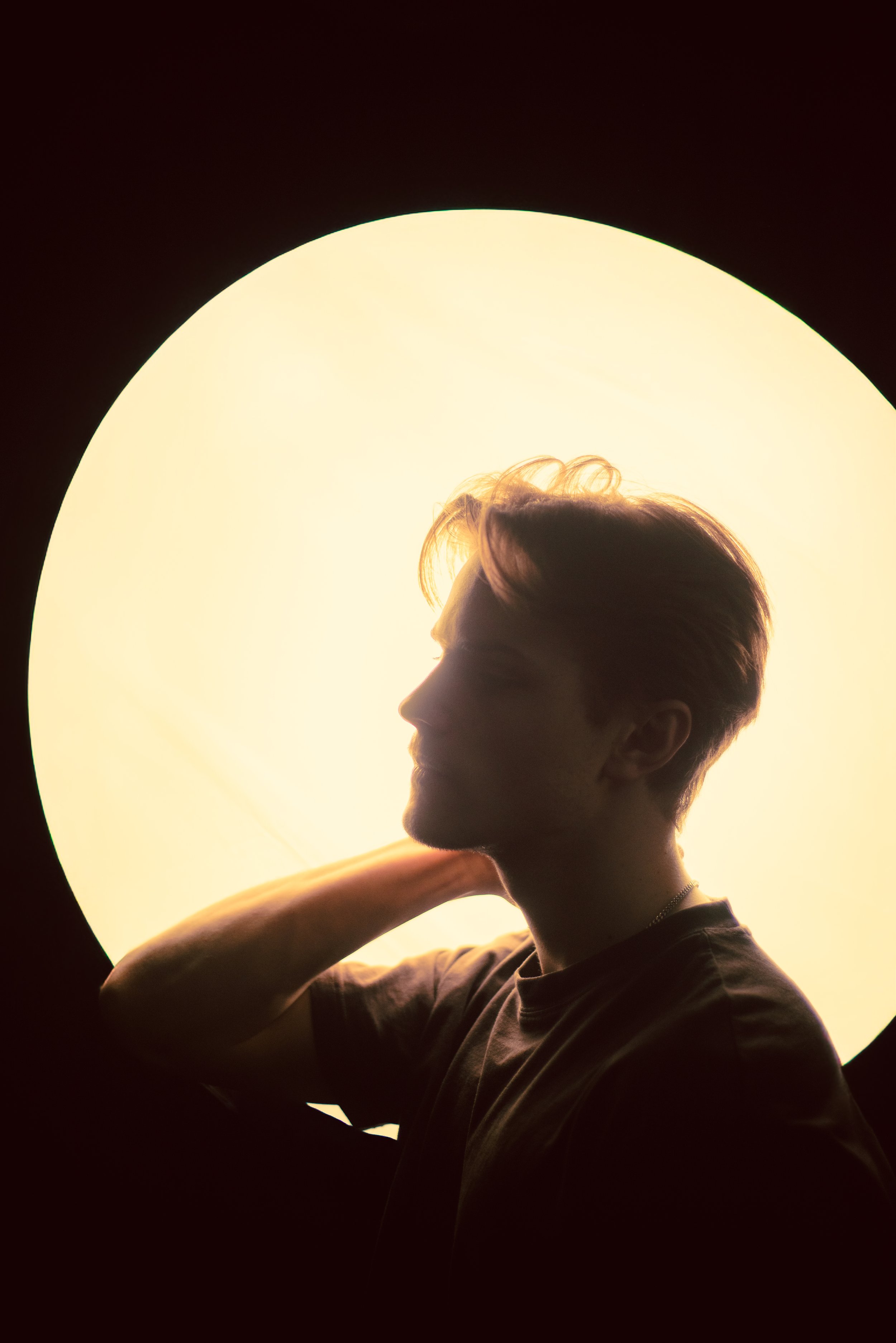 Profilbild av en person med kort hår som vilar sin hand på axeln framför en stor, varm ljuskälla.