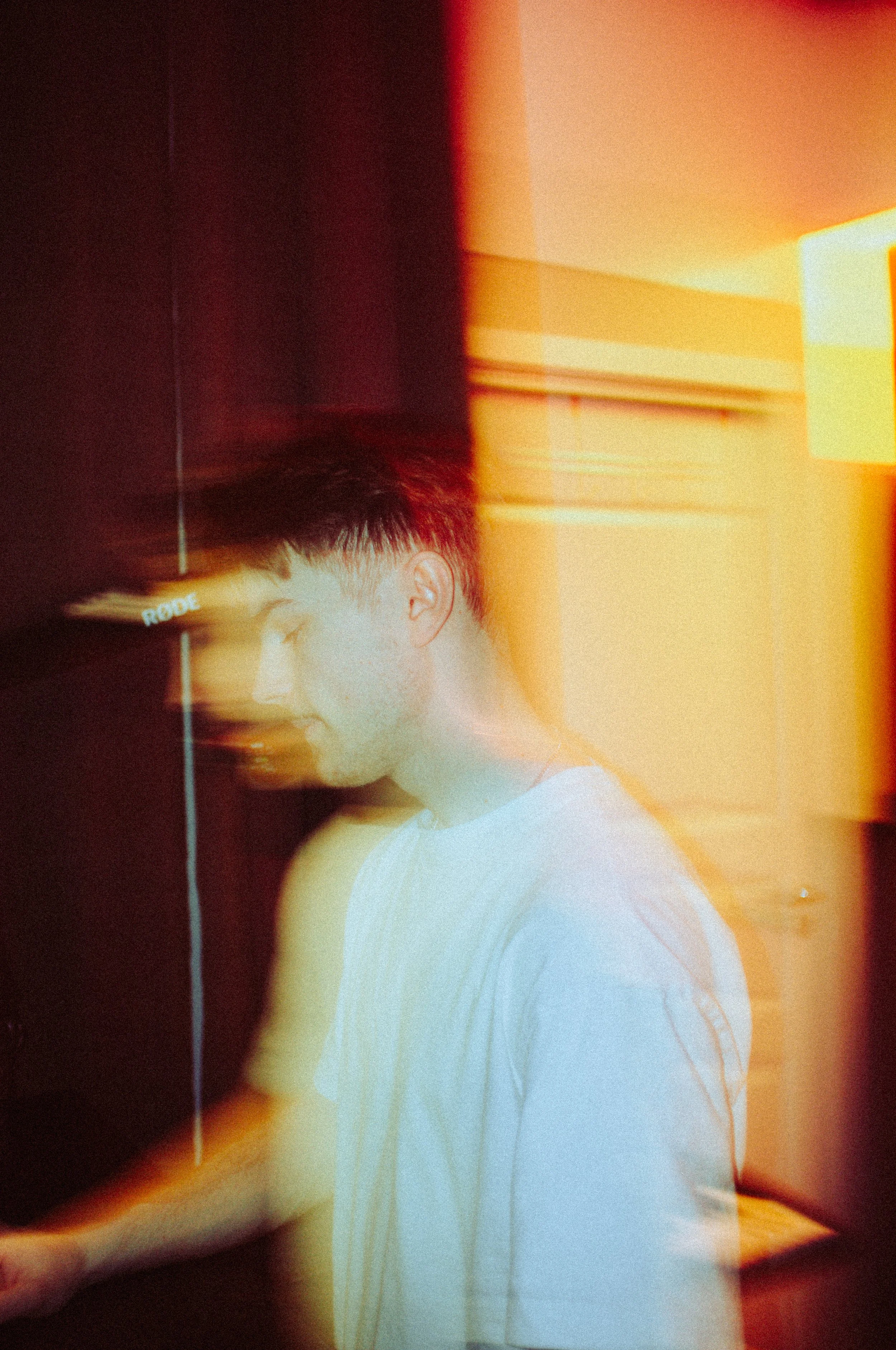 A person with short hair and a white sleeveless shirt standing indoors near a wall with warm lighting. The image has a layered, distorted, and abstract visual effect with color streaks.