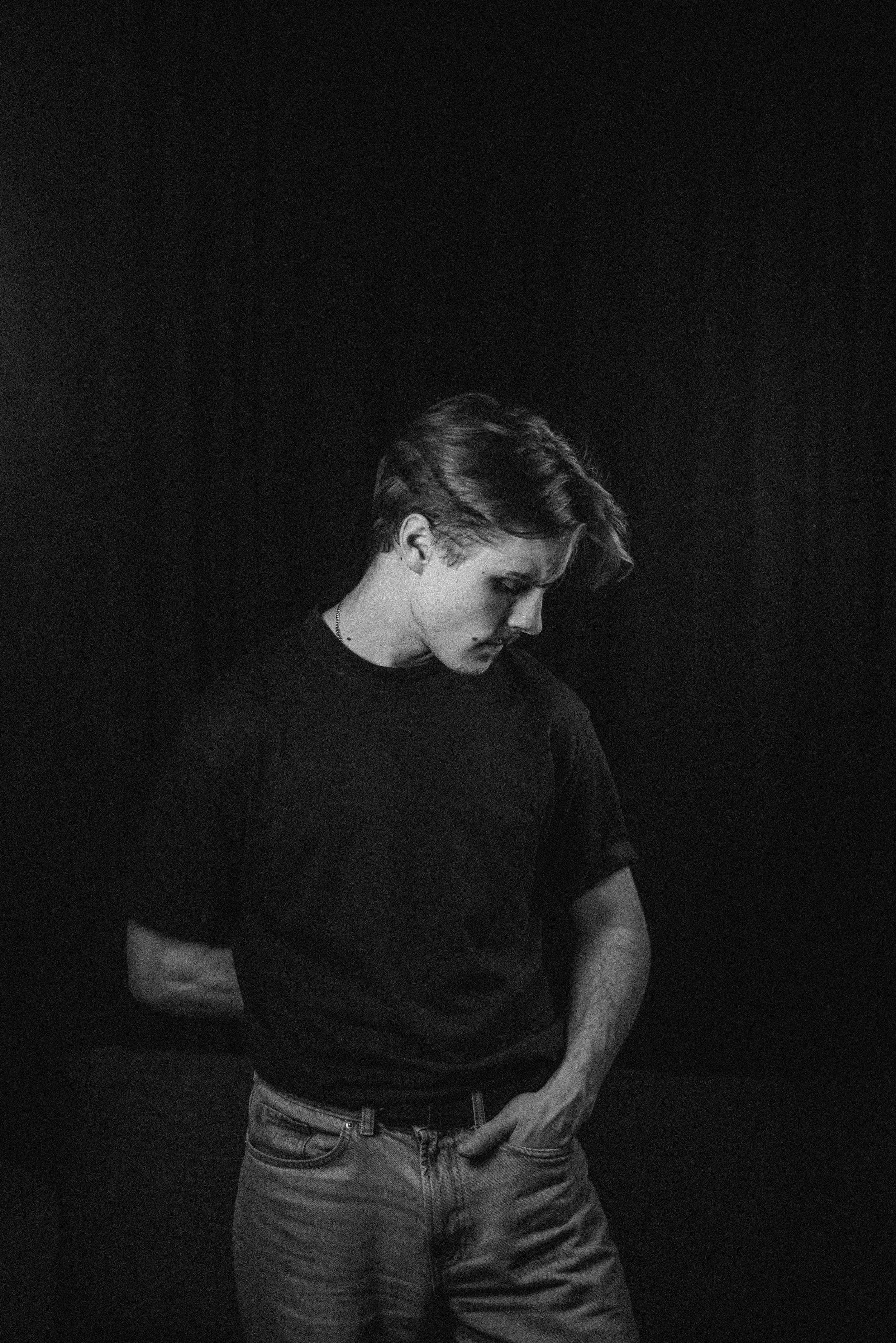 A young man with styled hair, wearing a black t-shirt and jeans, with his head slightly bowed and one hand in his pocket against a dark background.
