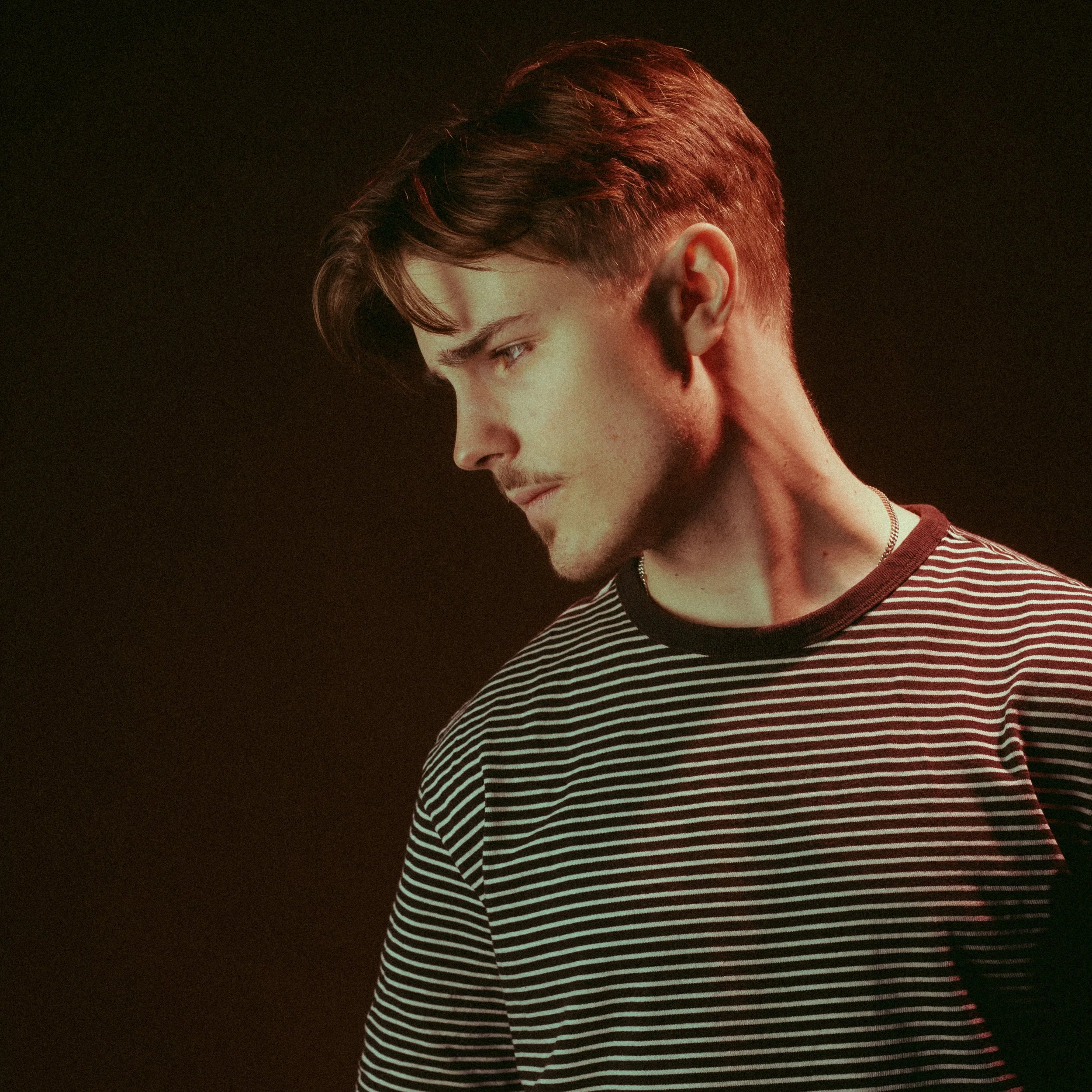 Close-up of a young man with short brown hair, wearing a black and white striped shirt, against a dark background.