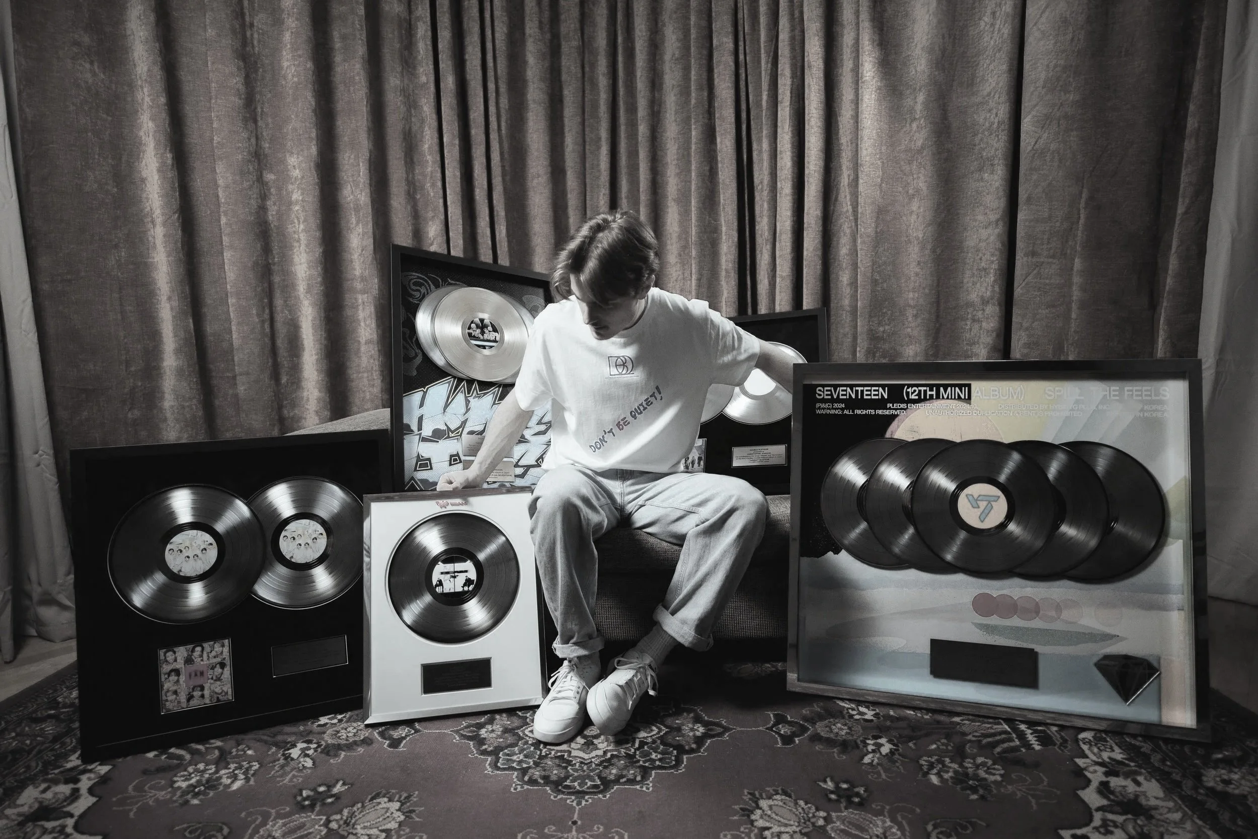En person som sitter bland flera vinylskivor och albumomslagsbilder på en soffa med mörka gardiner i bakgrunden.