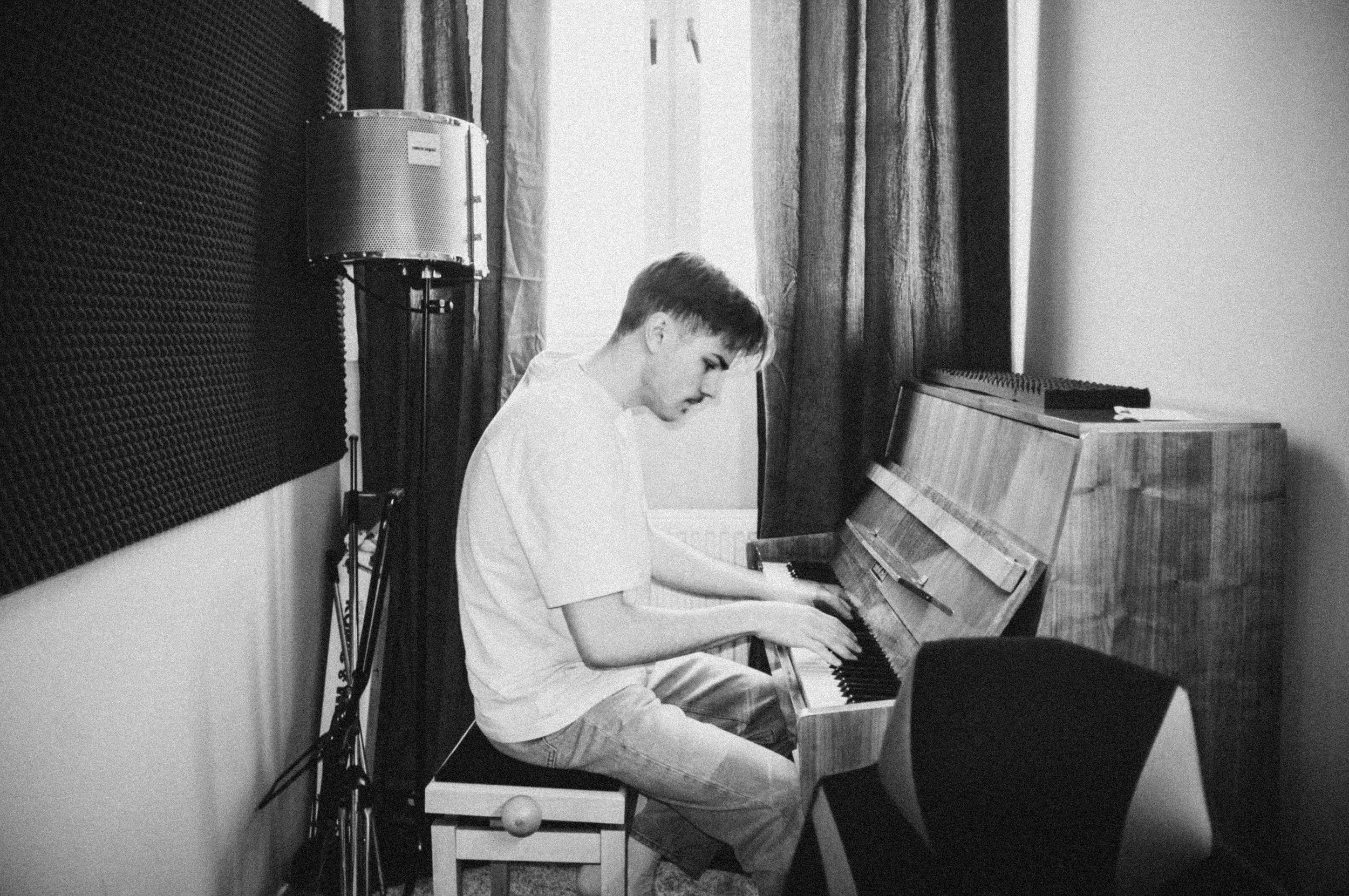 En ung man spelar piano i ett rum med ljudabsorberande väggpaneler och mörka gardiner framför ett fönster.