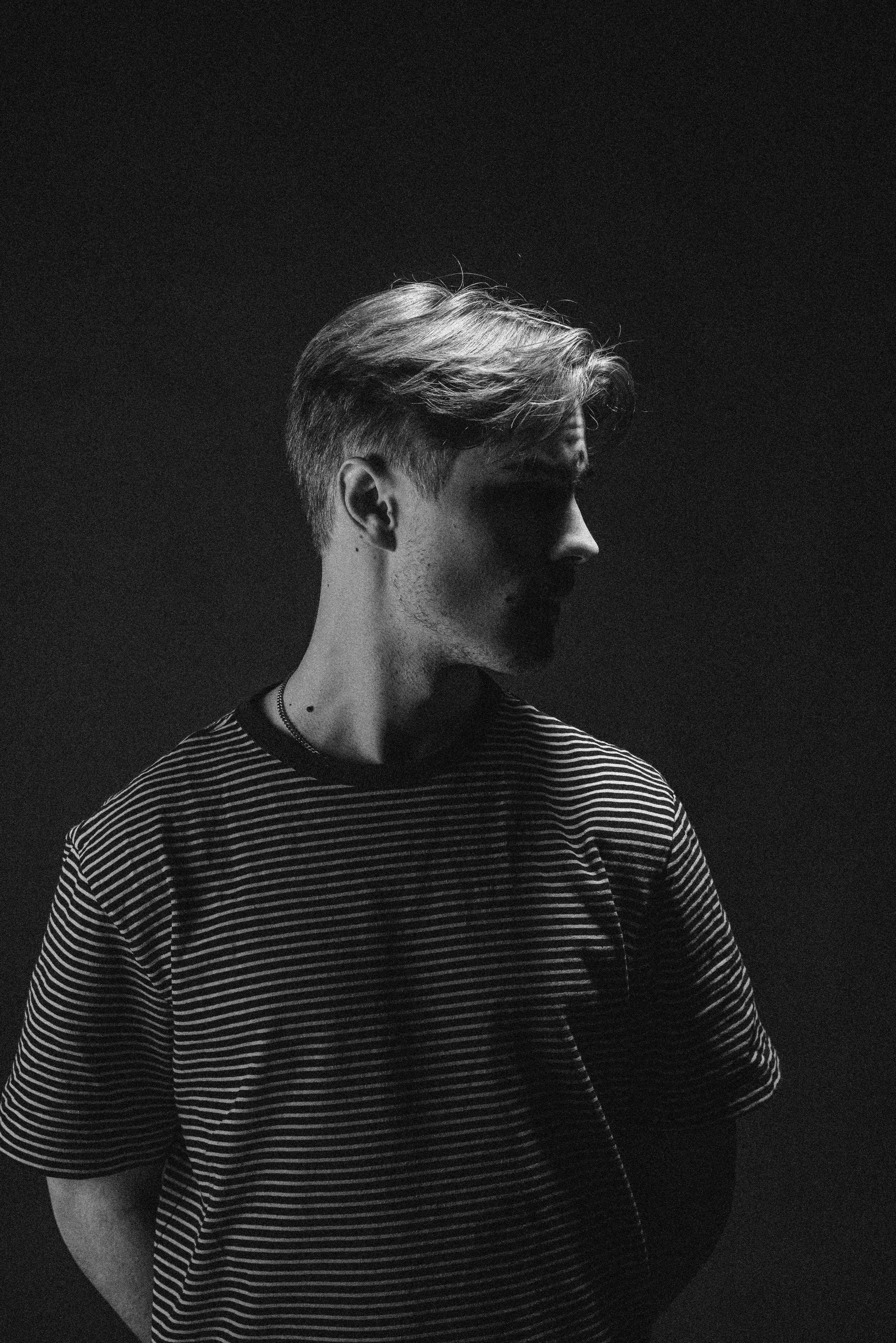 Black and white photo of a young person with short, styled hair, wearing a striped t-shirt and a necklace, standing against a dark background.