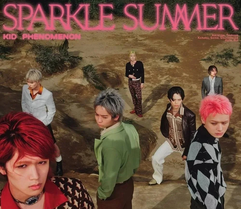 Gruppbild av KID PHENOMENON i olika kläder stående på en ökenliknande plats med stora stenar. Texten "Sparkle Summer" i rosa medelstora bokstäver och ytterligare text nedan i rosa.