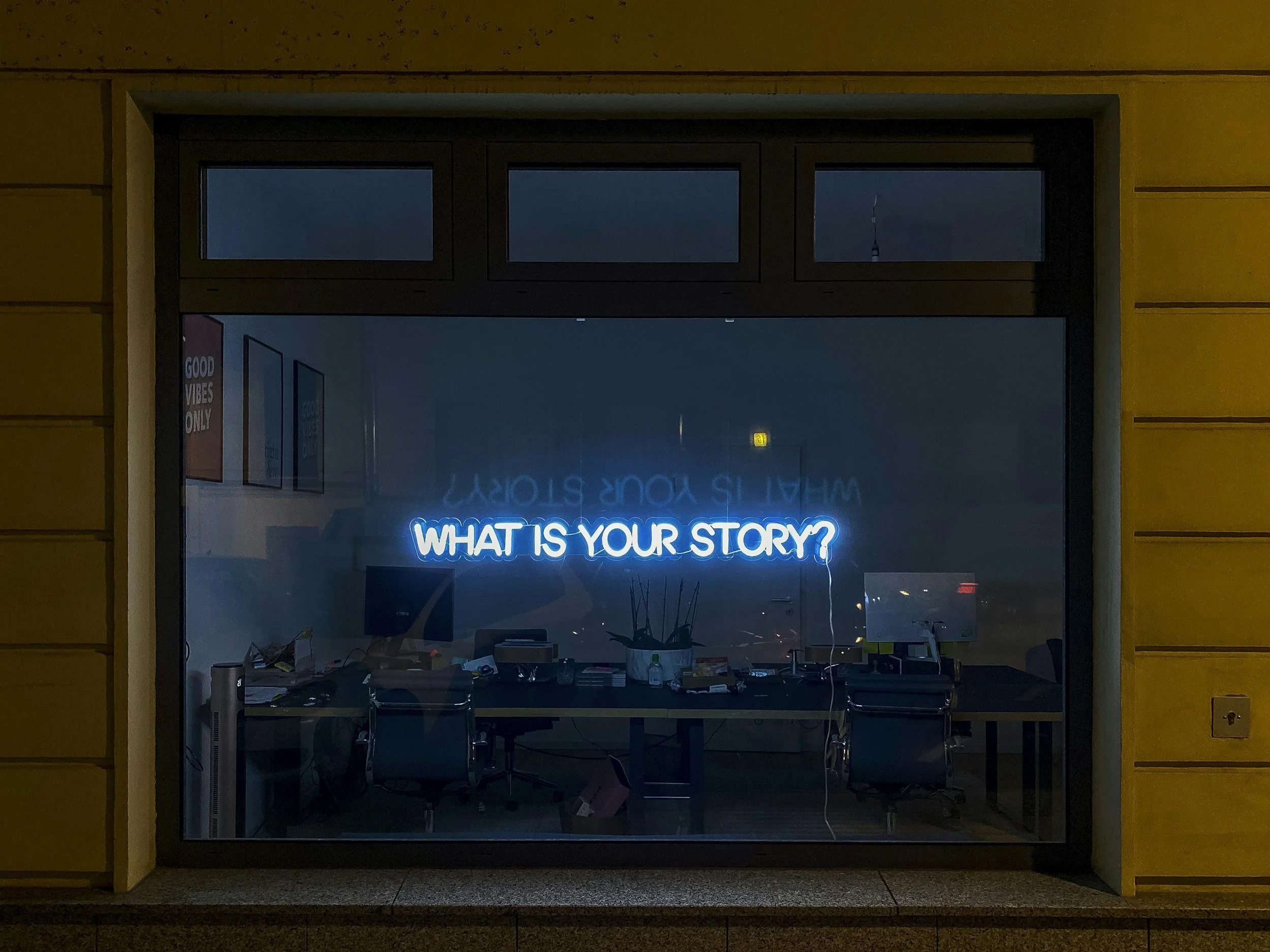 Nighttime view through a large window showing a brightly lit blue neon sign on the inside that reads, 'WHAT IS YOUR STORY?' The window frame is black, and the exterior wall is yellow with horizontal siding. Inside, there is a cluttered desk with various objects and a chair.