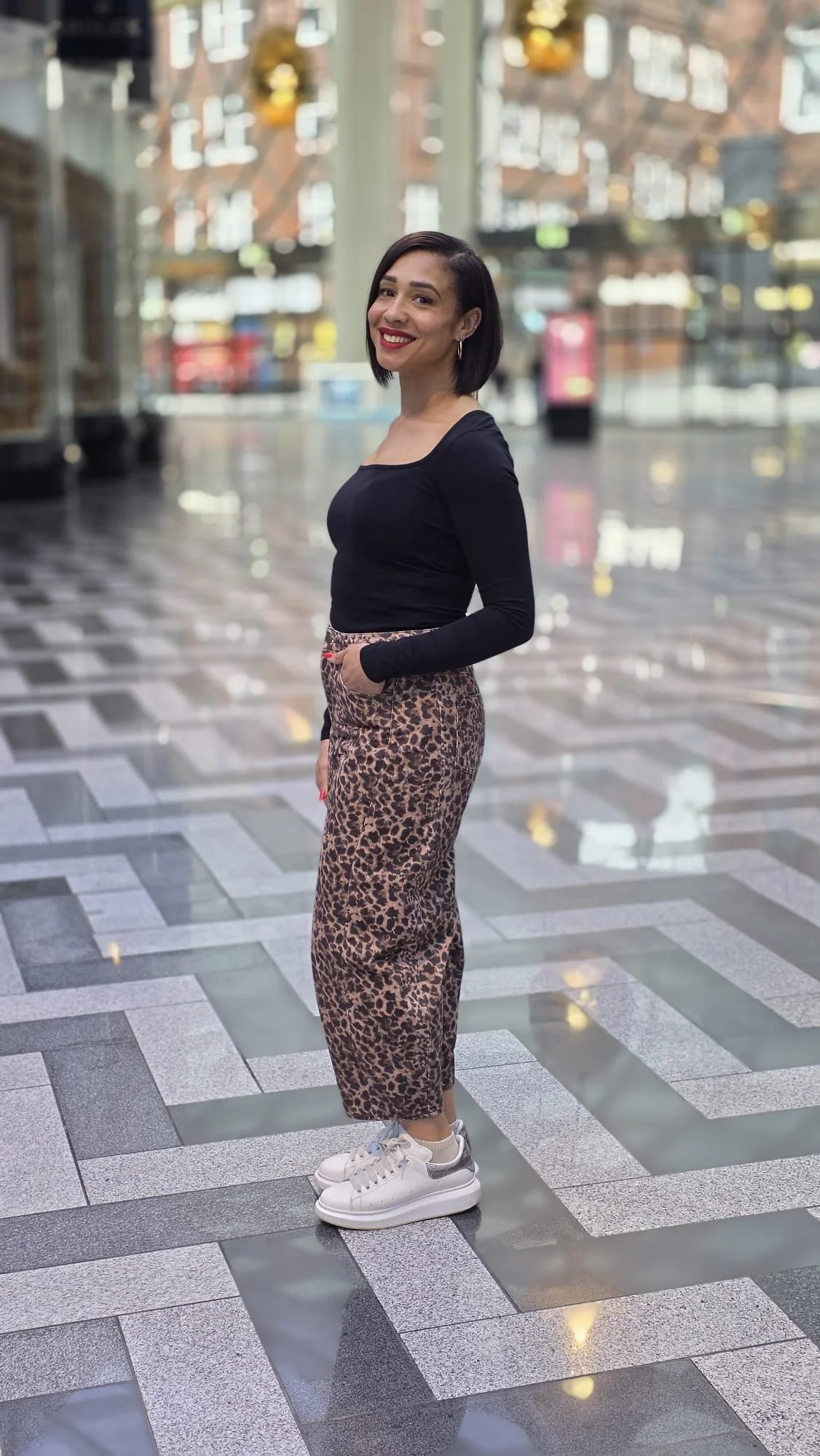 A woman with short black hair and red lipstick smiling, standing indoors with a glass exterior background, wearing a black long-sleeve top, leopard print pants, and white sneakers.