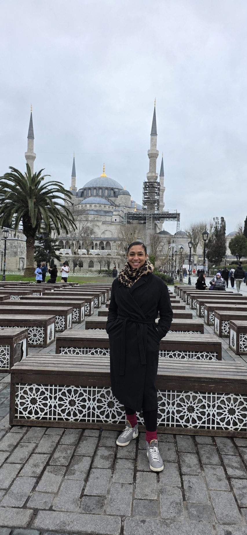 A woman standing and smiling in front of the Blue Mosque in Istanbul, Turkey, with benches, trees, and other visitors in the background under an overcast sky.