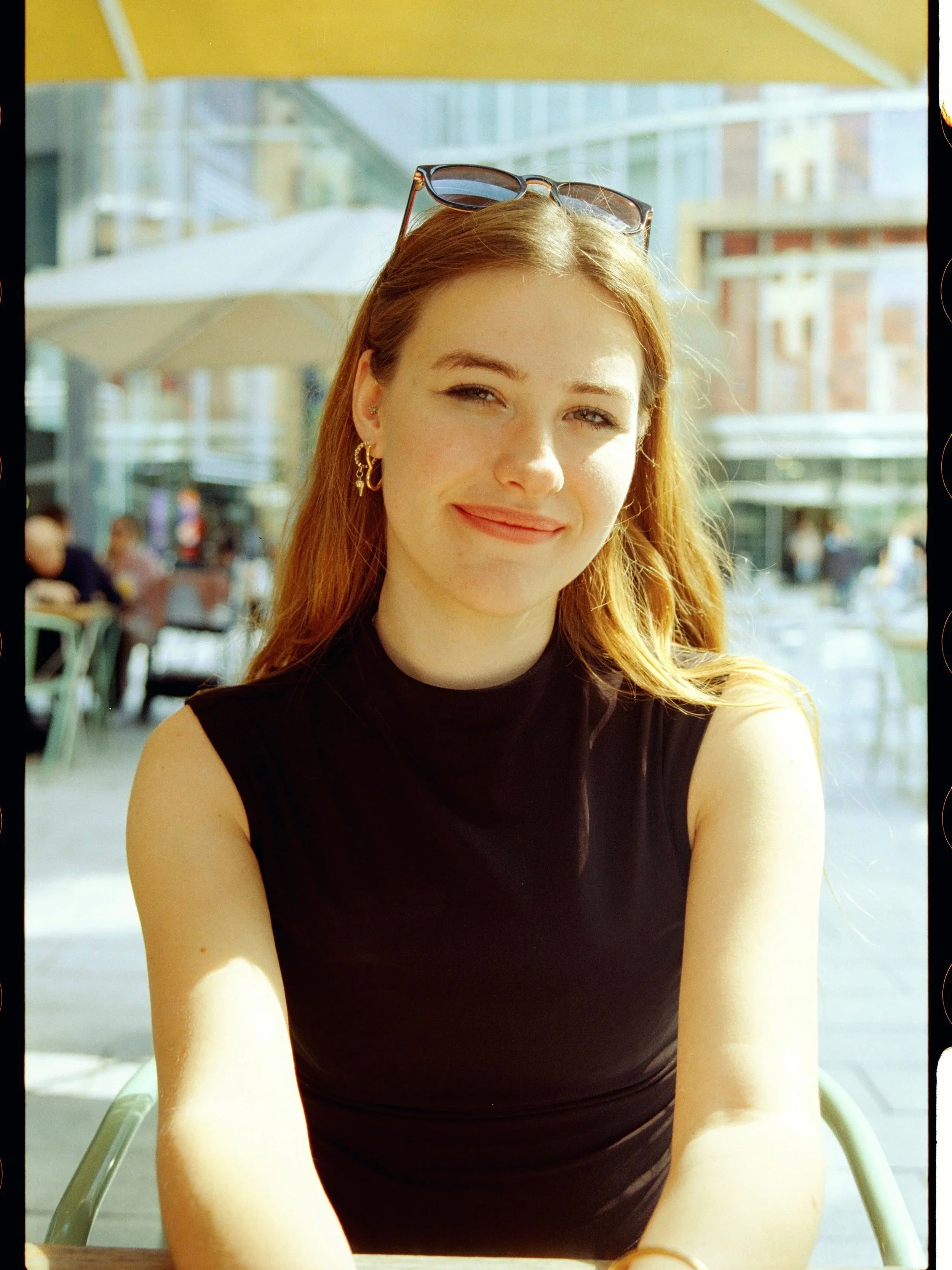 A young woman with red hair sitting outdoors at a table, wearing a black sleeveless top and sunglasses on her head, smiling.
