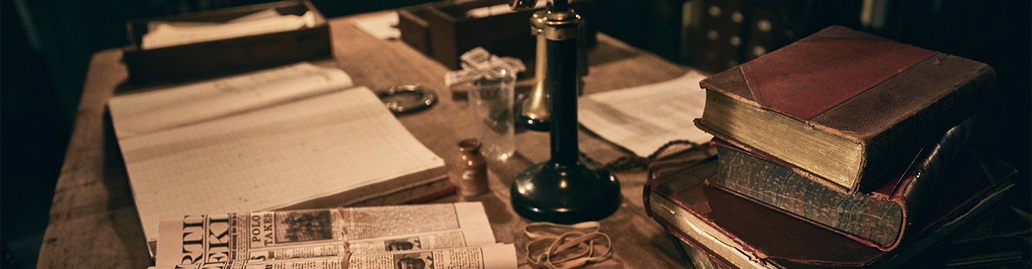A cluttered wooden desk with old books, newspapers, a candle lamp, and various papers and small items, suggesting a vintage or historical setting.