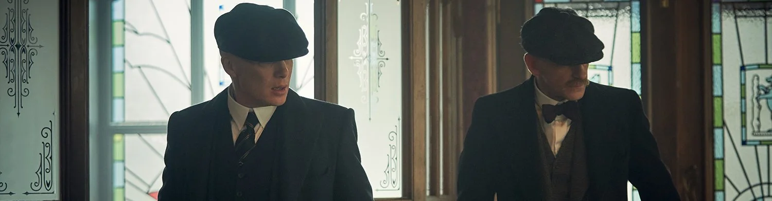 Two men dressed as early 20th-century gangsters with black hats, suits, and ties in a stained glass window-lit room.