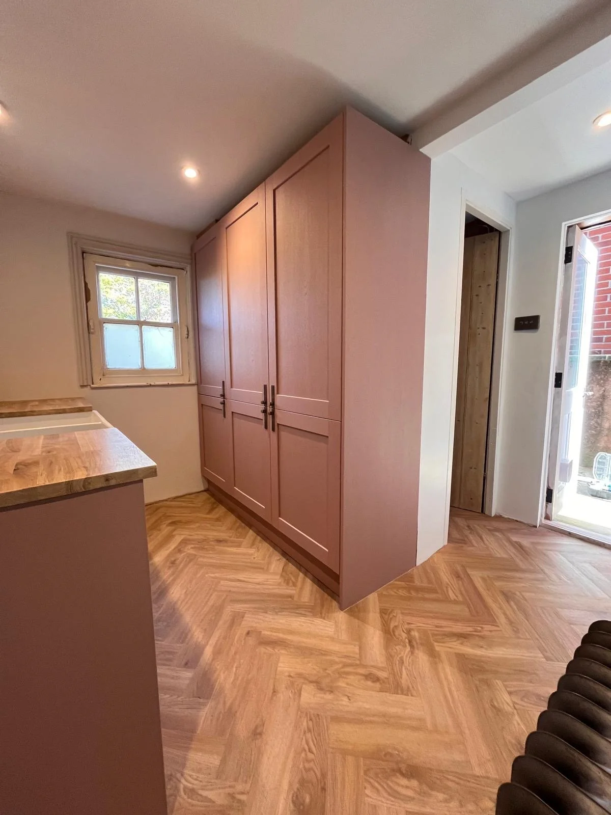 Interior of a kitchen with a large pink cabinet, a small window, wooden flooring, and a door leading outside.