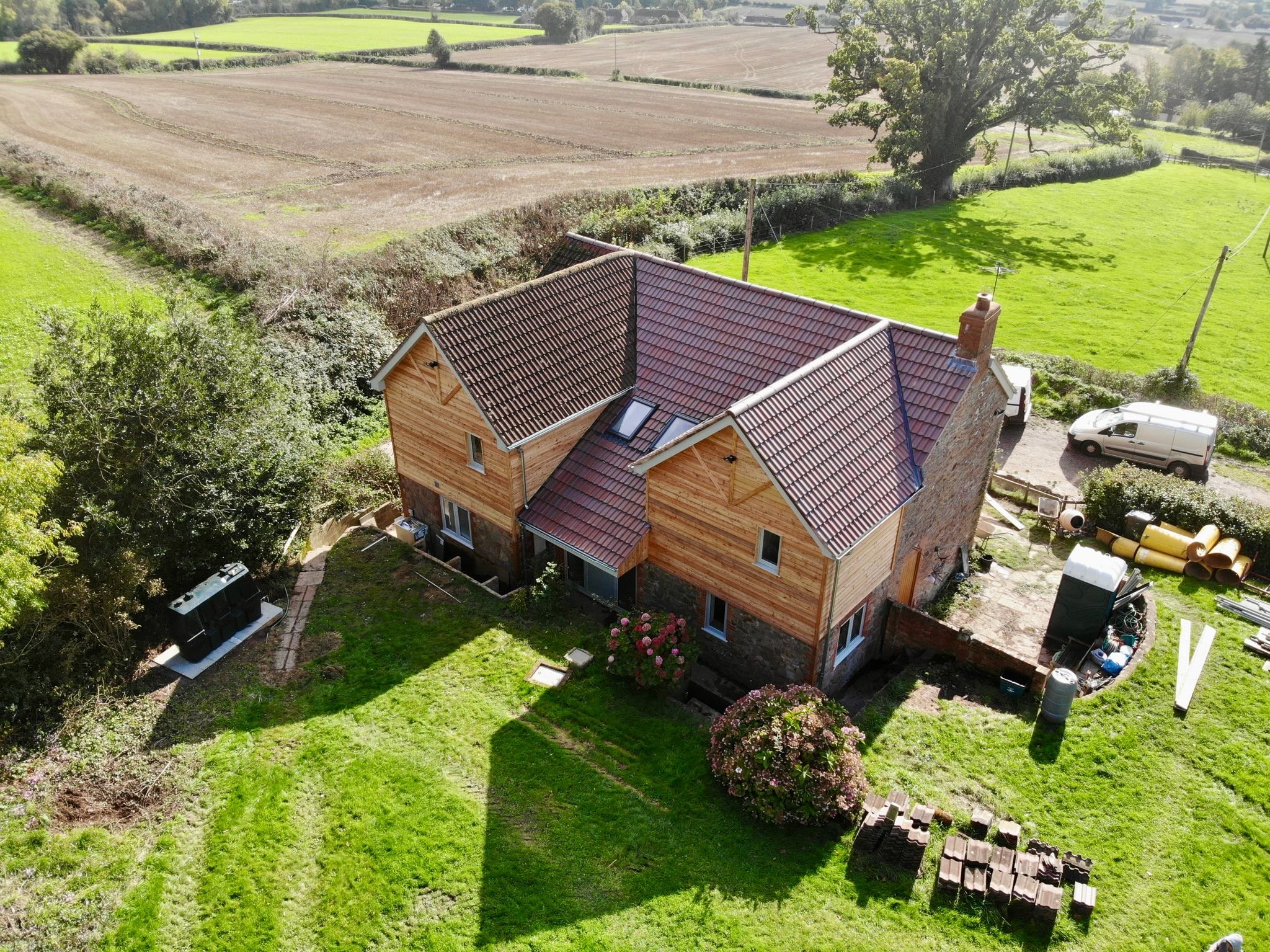 Aerial view of a house with a red tile roof and a brick chimney, surrounded by lush green grass, trees, and a garden. There is a white van parked in the driveway, and fields extend into the distance.