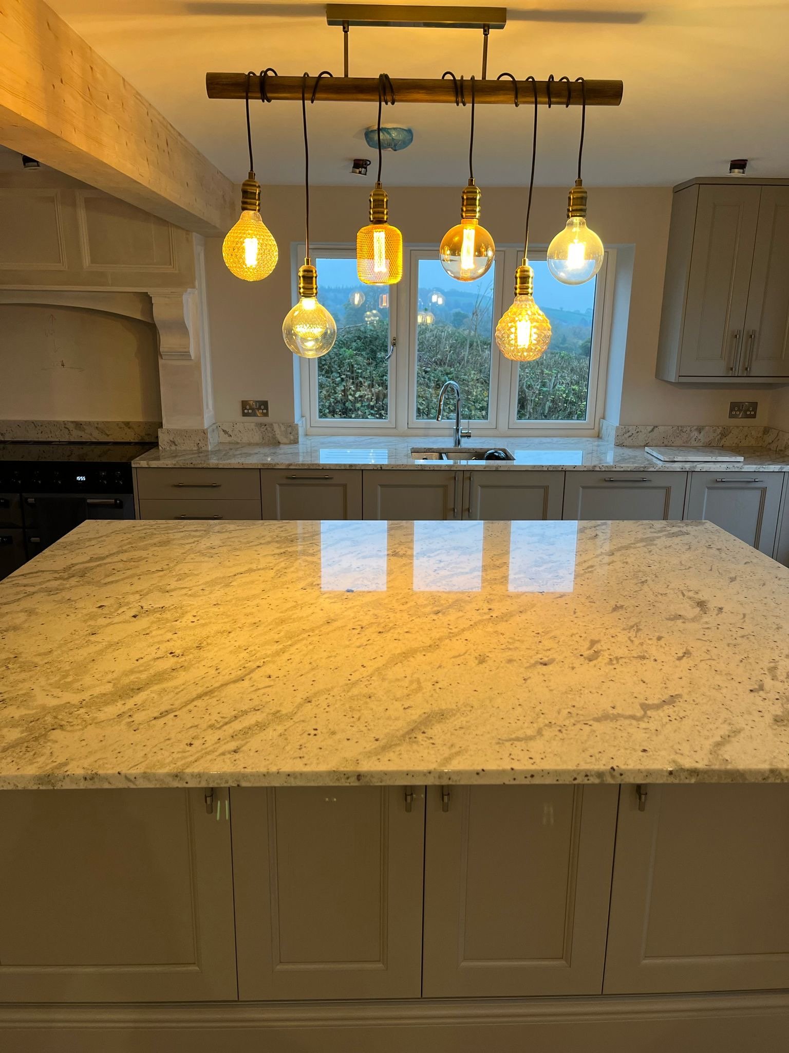 A kitchen with a large beige granite countertop, cream-colored cabinets, a window overlooking an outdoor landscape, and a wooden light fixture hanging from the ceiling with multiple exposed Edison bulbs.
