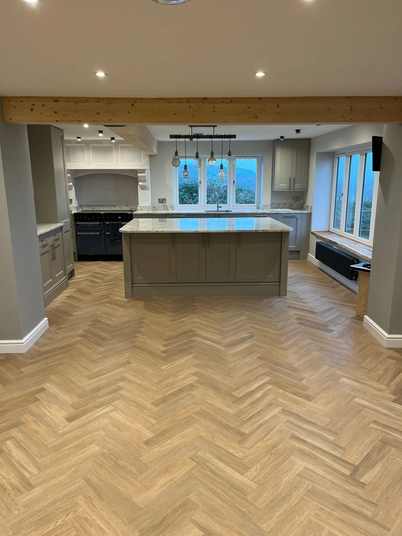 A modern kitchen with beige cabinets, a large marble island, light wood herringbone floor, three pendant lights hanging above the island, a window behind the sink showing a view of trees and mountains, and a radiator beneath the window.
