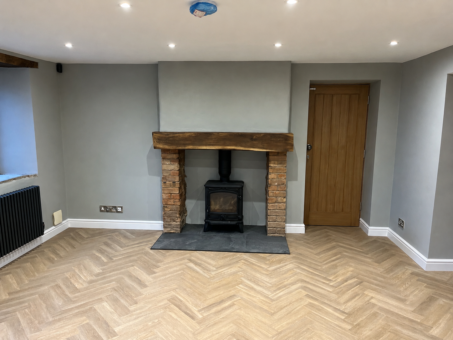 Living room with a brick and wood fireplace, light grey walls, wood floor, and a brown wooden door.