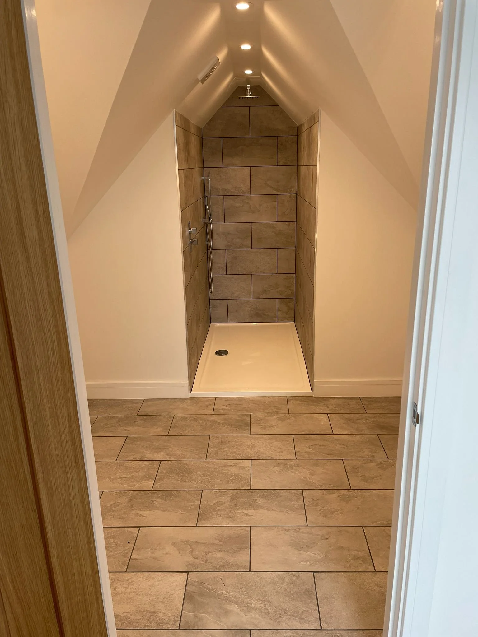 A modern walk-in shower with beige tiled walls and matching floor tiles. The ceiling is sloped with recessed lighting, and the shower has a handheld showerhead with a wall-mounted control. The shower drain is visible on the floor.