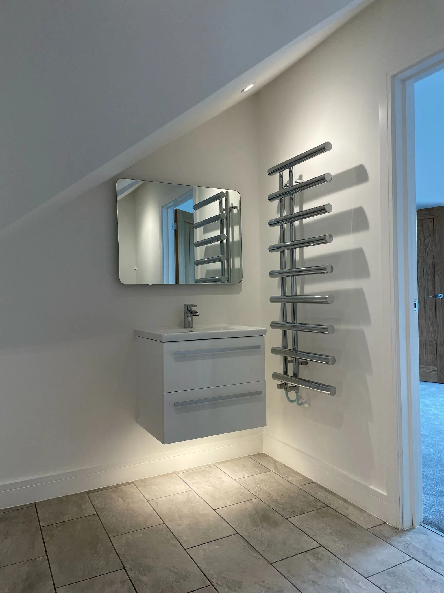 A bathroom with a white wall-mounted sink with a faucet, a mirrored medicine cabinet above, a modern silver towel rack on the wall, a window on the right, and tile flooring.
