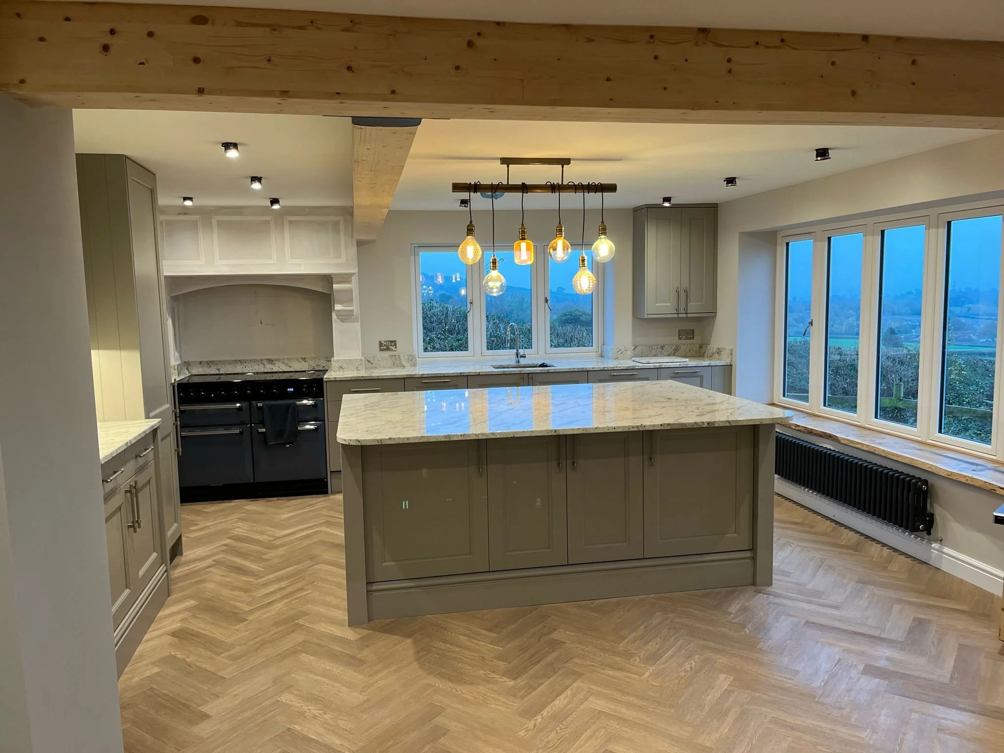 Modern kitchen with an island, pendant light bulbs, large windows, and wood flooring.