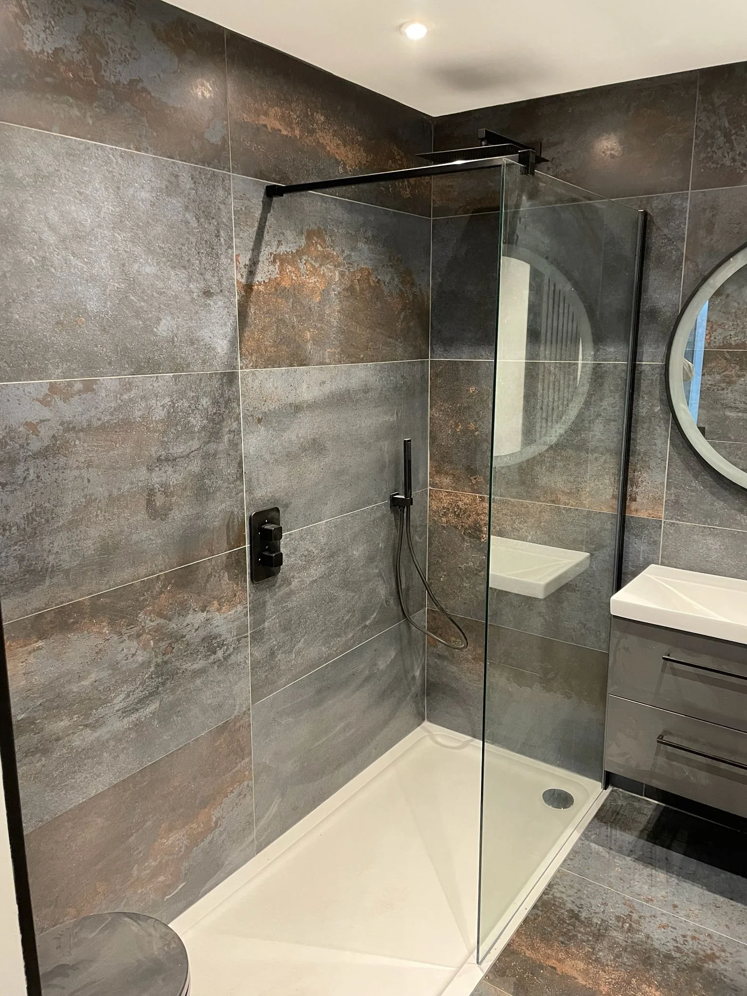 Modern bathroom with gray stone tiled walls, walk-in shower with glass partition, black shower fixtures, small white shower base, oval mirror, and black vanity with white sink.