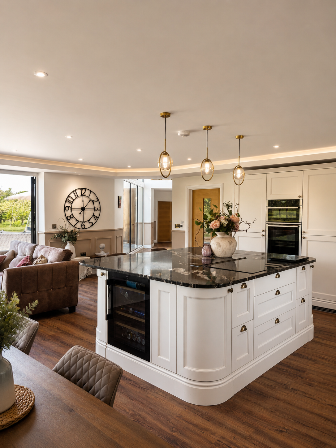 Open-concept kitchen with a marble island, pendant lights, white cabinetry, a built-in oven, and a floral centerpiece, adjacent to a living room with a large wall clock, a brown sofa, and large windows showing greenery outside.