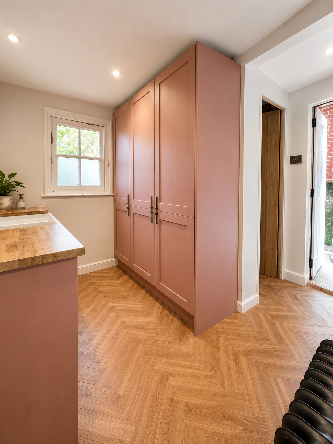 A kitchen corner with pink cabinets, a white window, wooden countertop, hardwood floor, and a door leading outdoors.