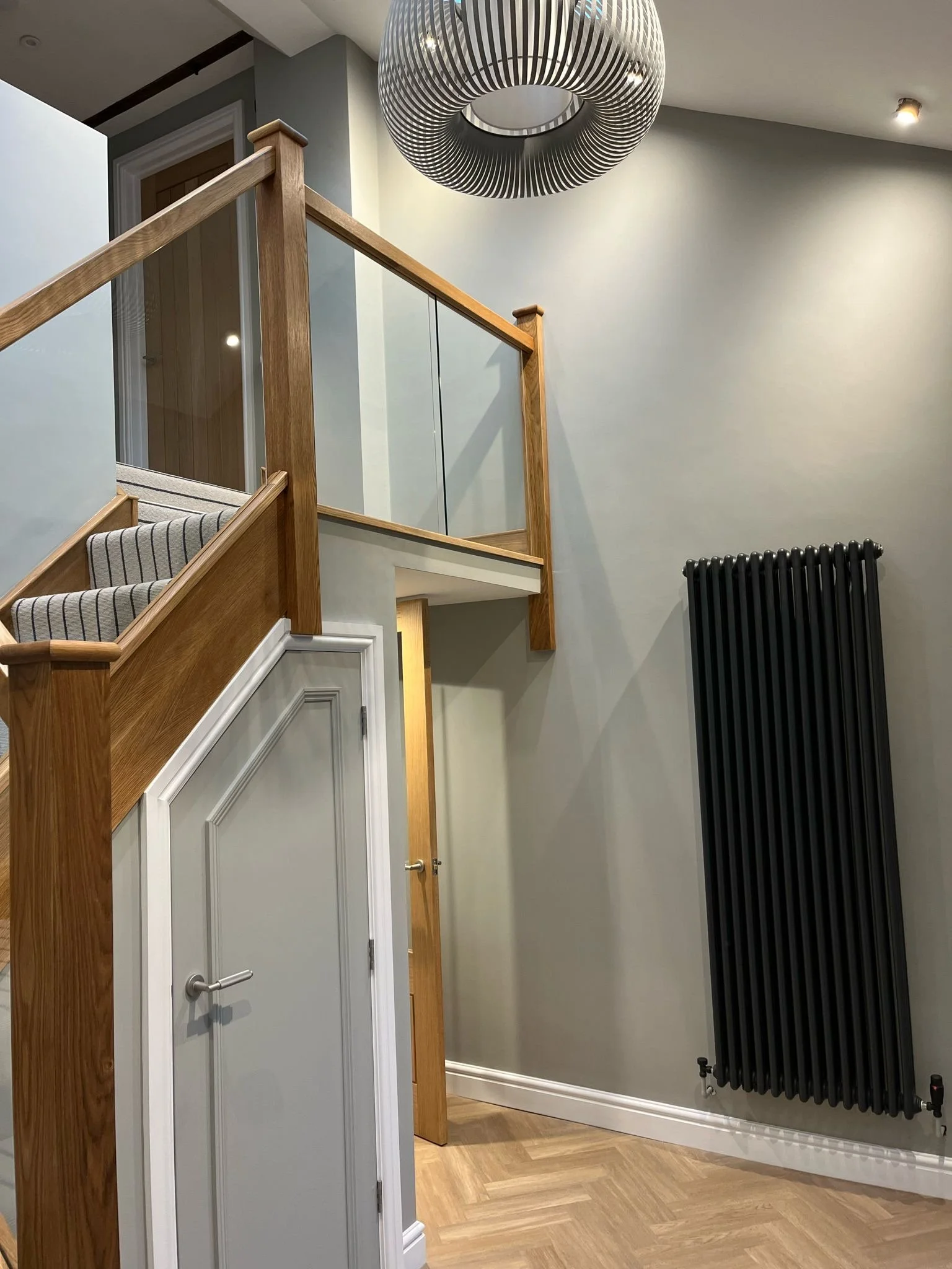 Interior view of a modern house showing a staircase with wooden handrails, a gray wall with a white door, a black radiator, and a circular hanging light fixture.