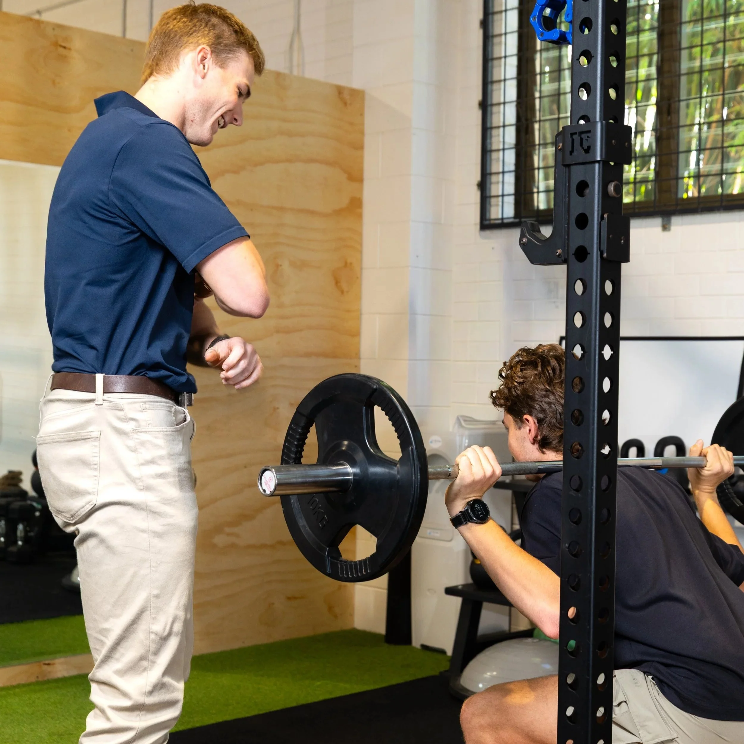 A sports physiotherapist coaching a client through a barbell squat exercise to optimise movement patterns and lifting mechanics