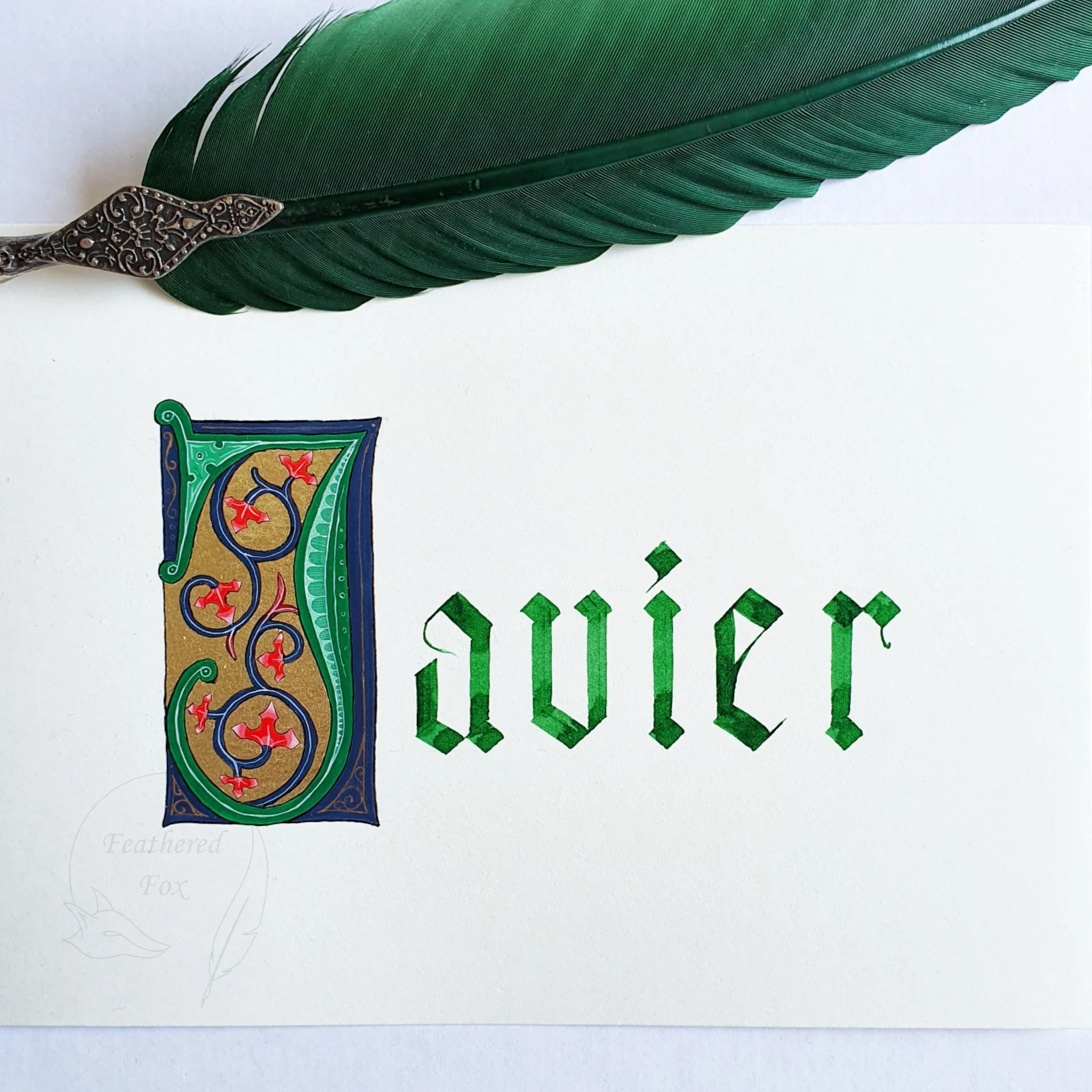 Un prénom calligraphié en vert dans un style gothique avec une majuscule enluminée et décorée de feuille d'or, avec une plume d'écriture verte posée au dessus.