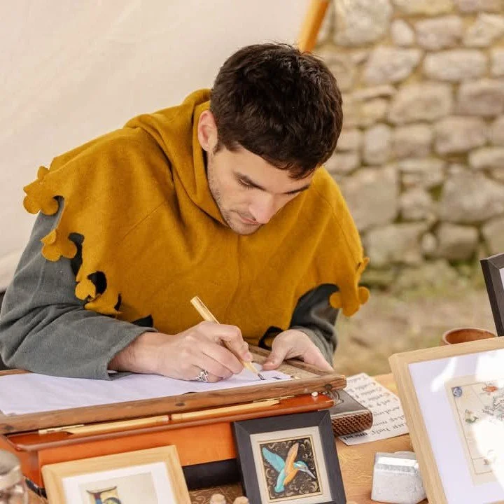 Un homme habillé d'une tunique médiévale avec un capuchon orange, assis à une table lors d'une fête médiévale pour calligraphier un texte. La table affiche des documents calligraphiés et enluminés et des objets liés au travail de scribe au moyen-âge.