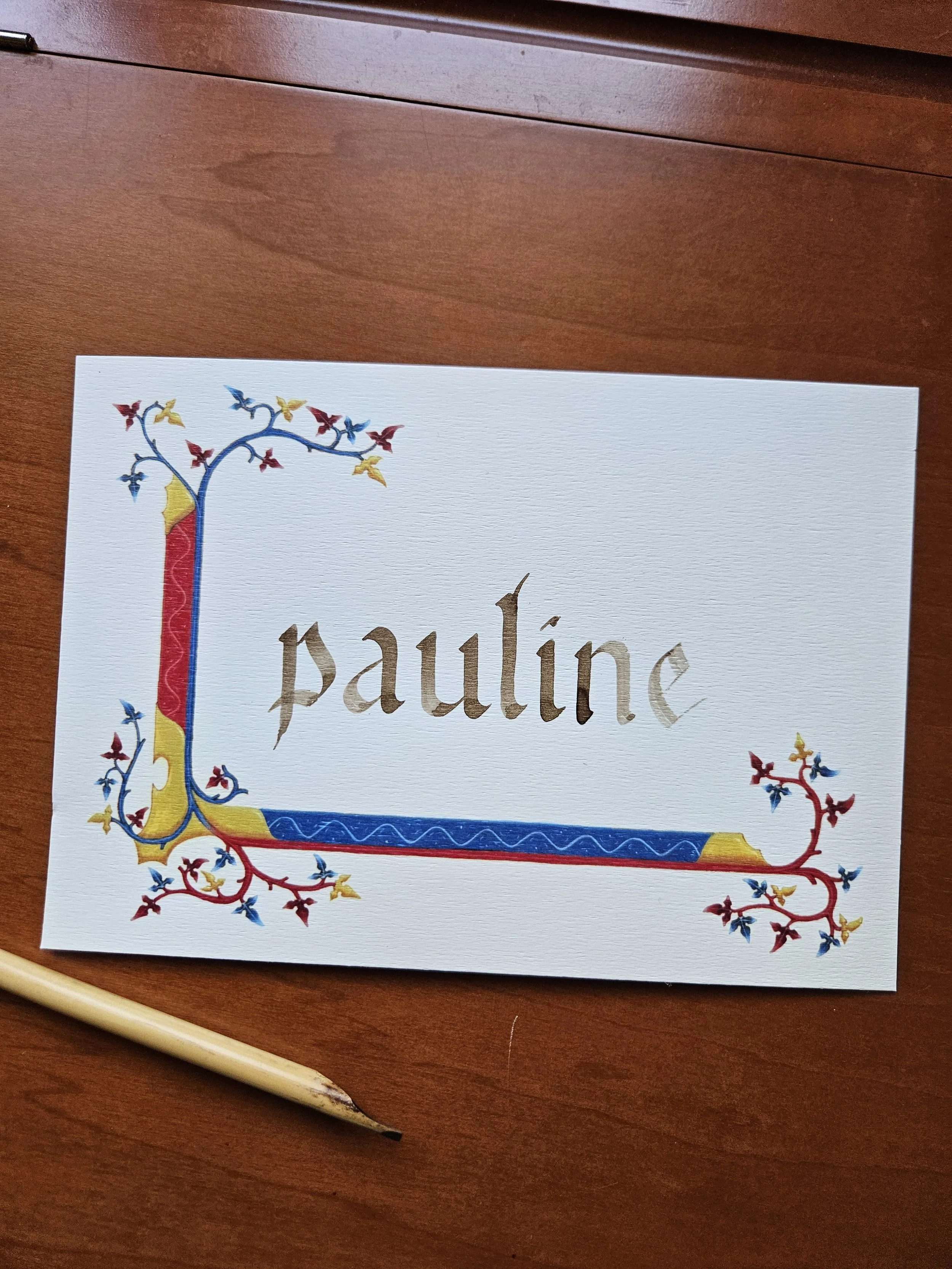Une carte avec le prénom 'Pauline' écrit en lettres brunes de style gothique, décorée de motifs enluminés en rouge, or et bleu, avec un calame posé au coin inférieur gauche.
