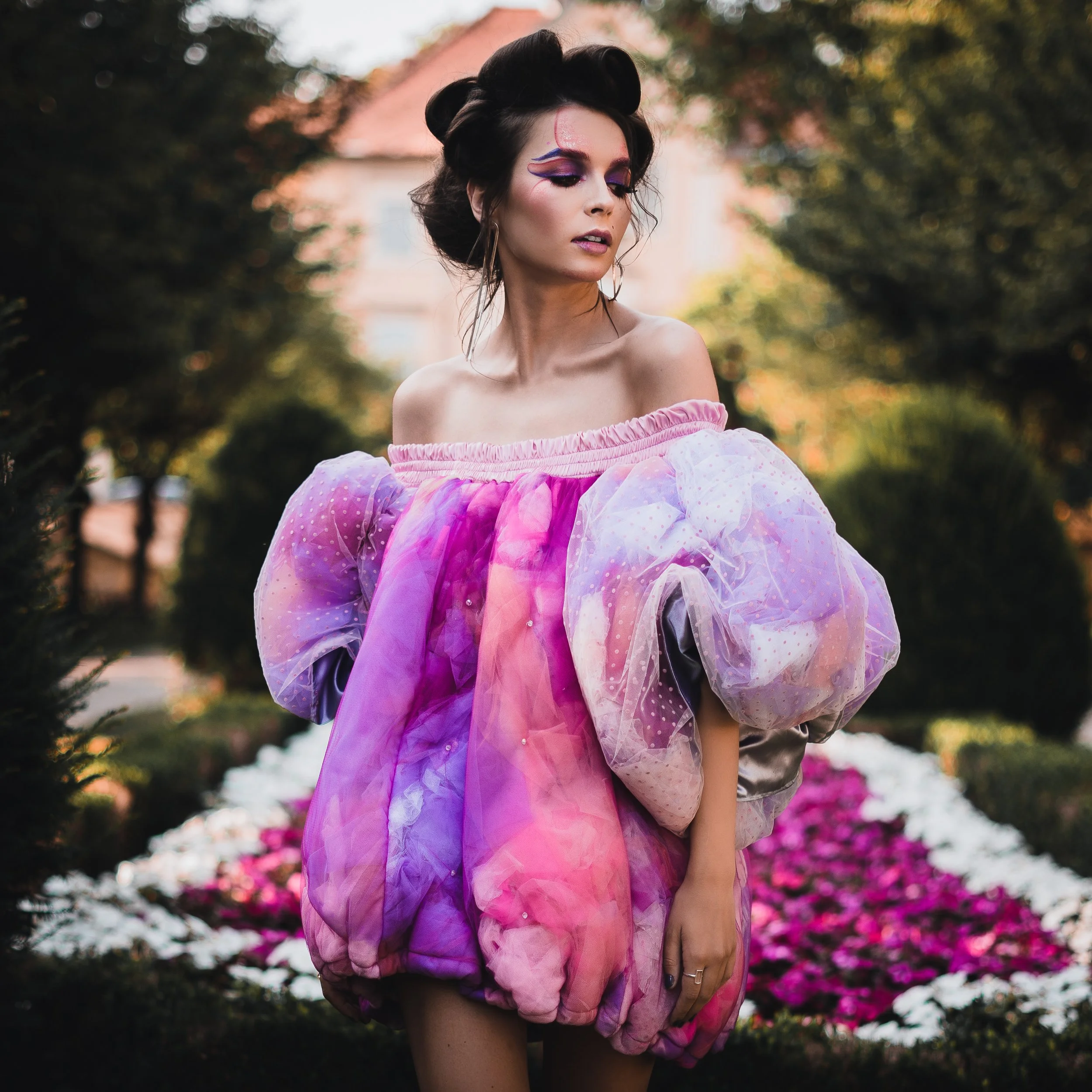 Eine junge Frau mit dunklen, hochgesteckten Haaren trägt ein rosa Kleid mit Tüll und Pailletten und posiert in einem Garten mit blühenden Blumen im Hintergrund.