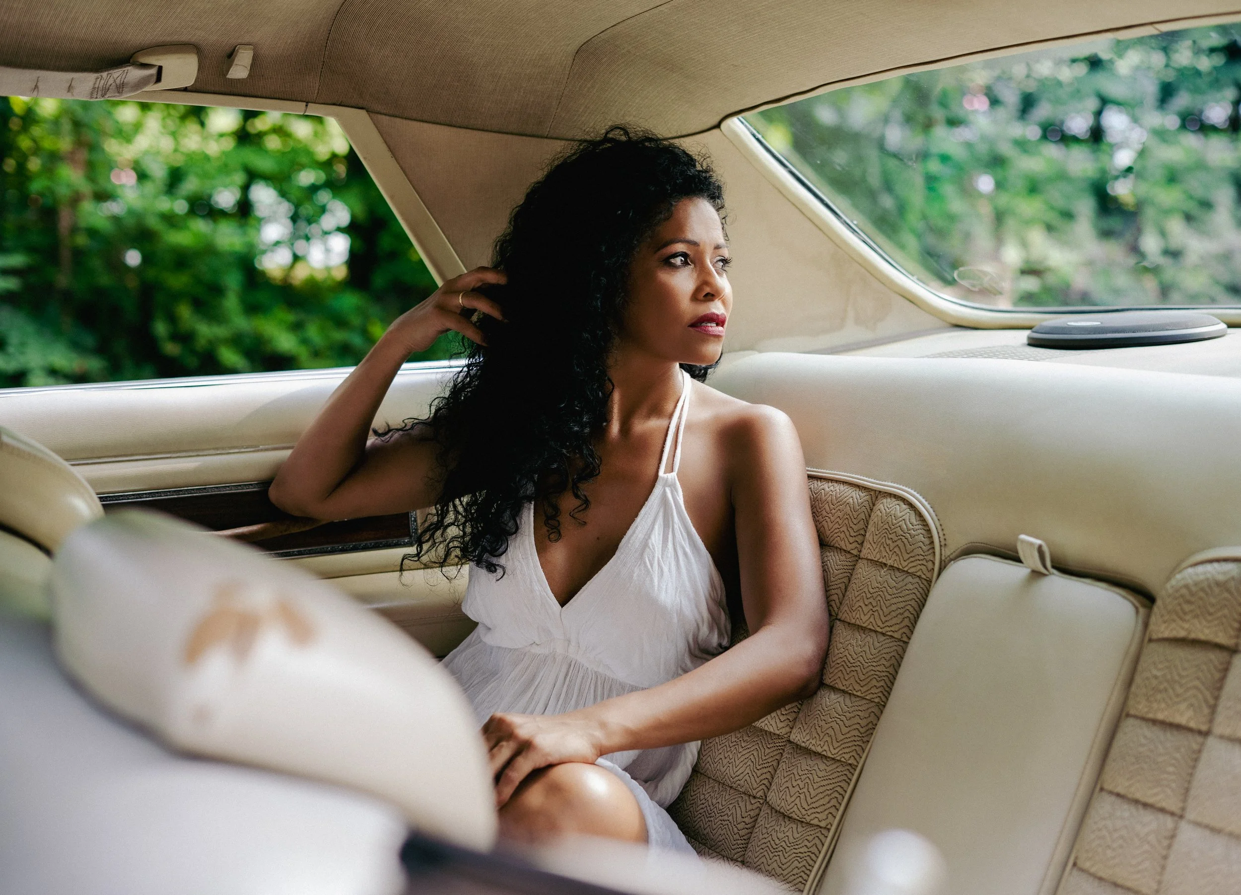 Eine junge Frau mit schwarzen, lockigen Haaren sitzt in einem Vintage-Auto. Sie trägt ein weißes, ärmelloses Kleid und blickt nach außen, während sie eine Hand an den Kopf hebt. Der Hintergrund zeigt grüne Bäume, was auf eine Fahrt durch die Natur hi