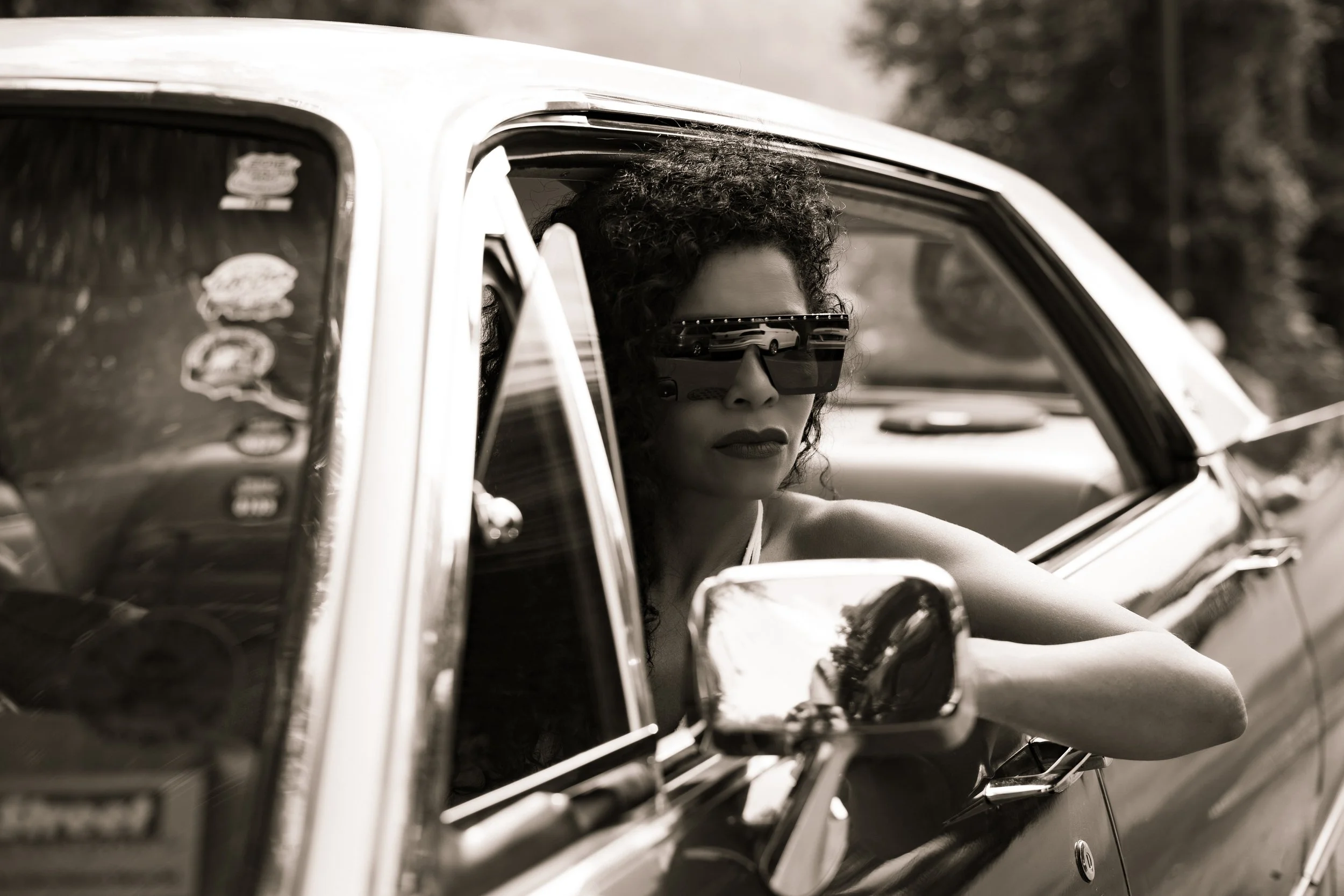 Nahaufnahme einer Frau mit lockigem Haar und Sonnenbrille, die aus einem Auto schaut, bei Schwarz-Weiß-Fotografie.